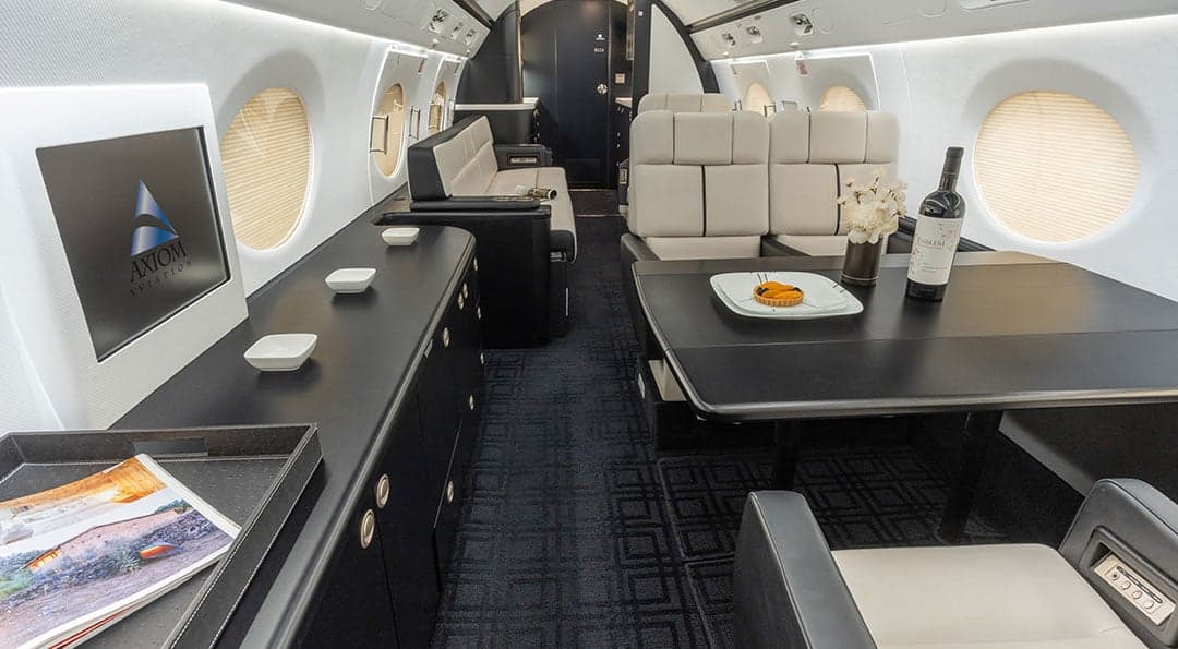 Interior view of the Gulfstream G550 cabin with a table, seats, and Axiom Aviation logo on a screen.