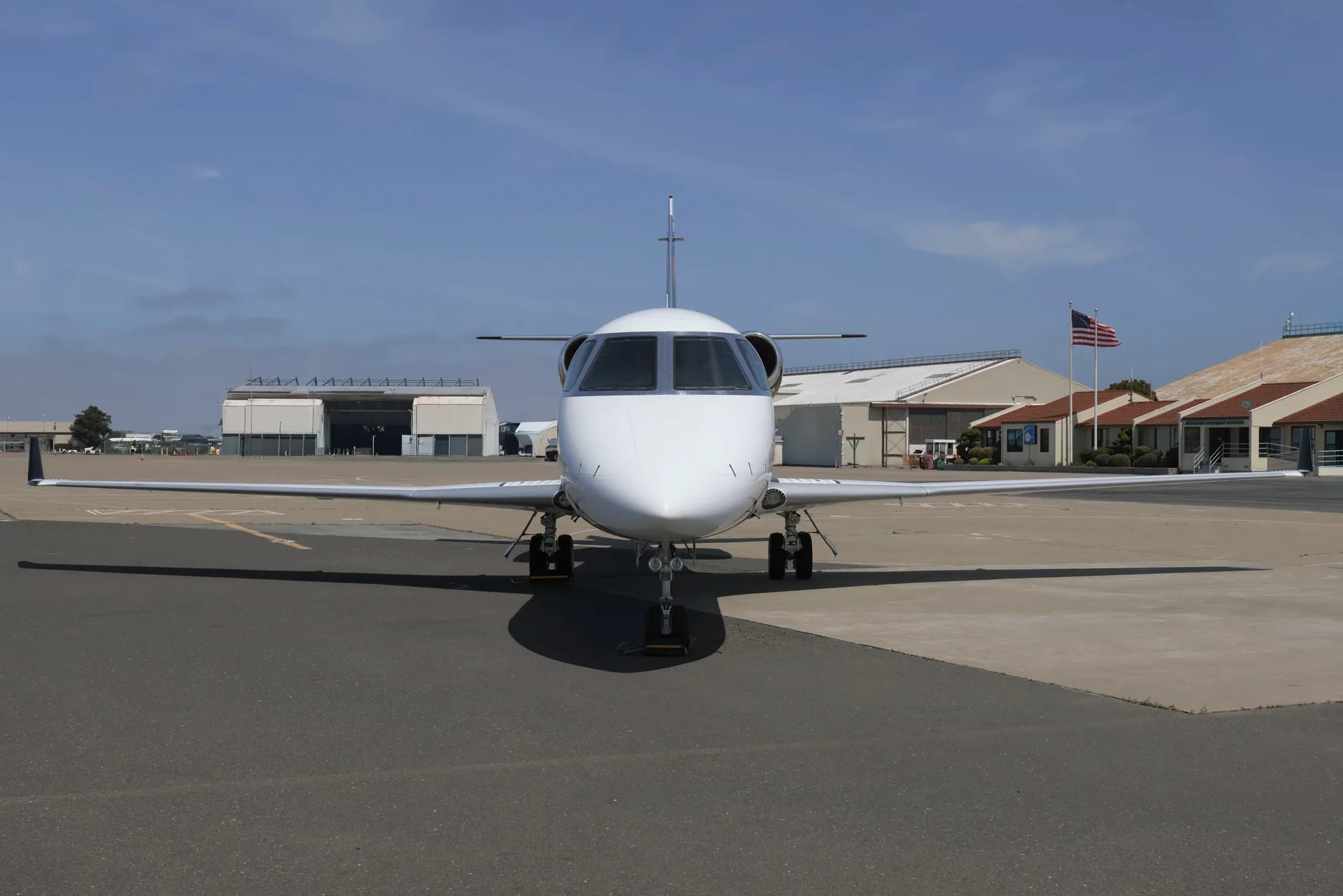 A front view of a white Gulfstream G280 business jet parked on an airfield with hangars in the background.