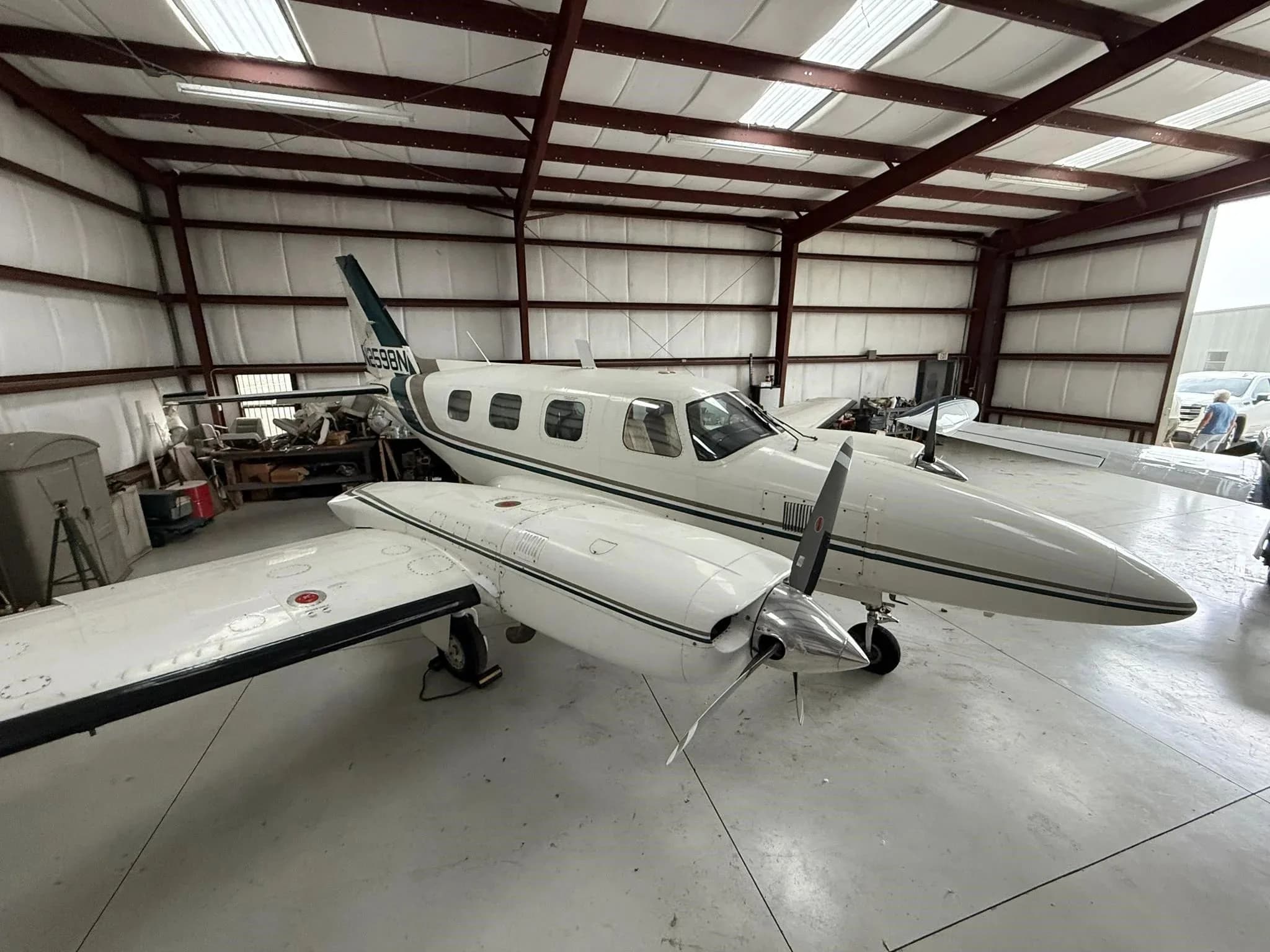 A white twin-engine Piper Navajo aircraft with green and brown stripes, seen from the front-right in a hangar.