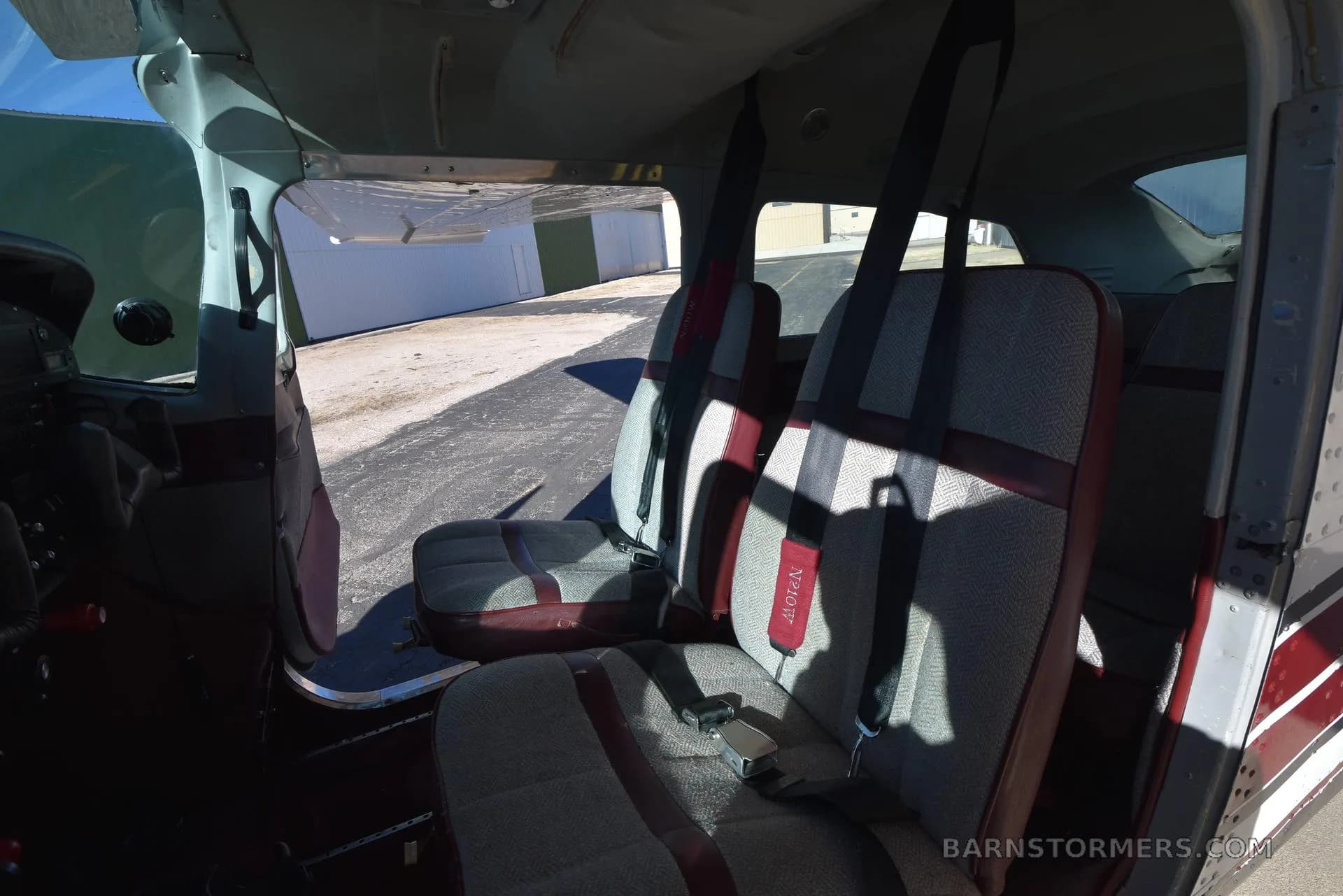 The interior cabin of a Cessna aircraft, showing two rows of grey and maroon fabric seats with seatbelts.