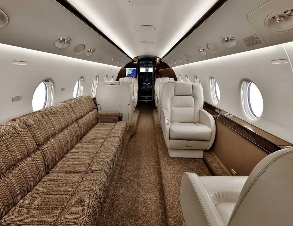 A view of the private jet's cabin, showing a long patterned sofa on the left and individual beige seats on the right.