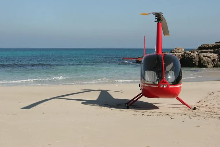 A red Robinson R44 helicopter parked on a sandy beach.