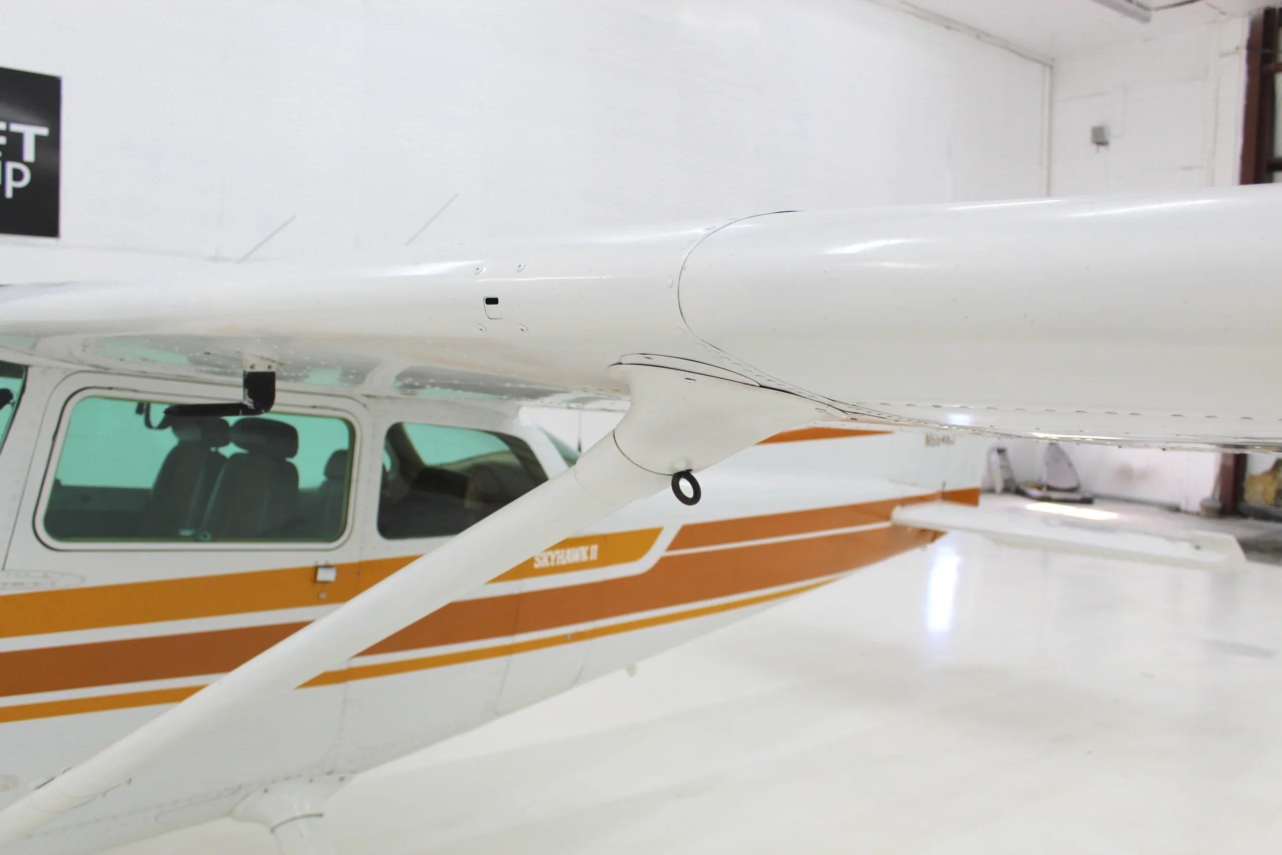 A close-up view of the wing strut and fuselage connection of a white and orange Cessna Skyhawk II in a hangar.