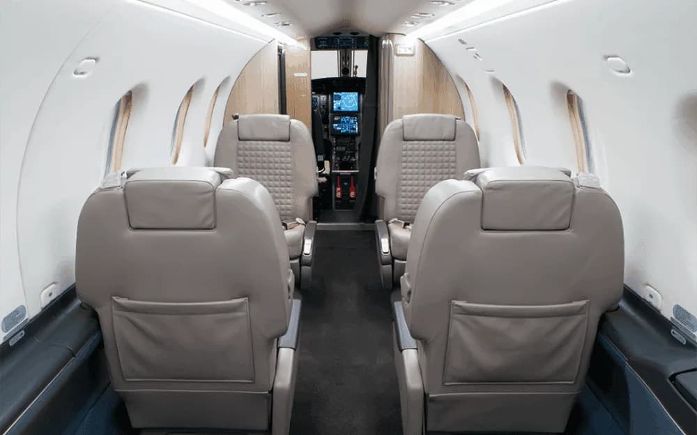 A view down the aircraft cabin aisle shows light grey leather seats and a glimpse of the cockpit beyond.