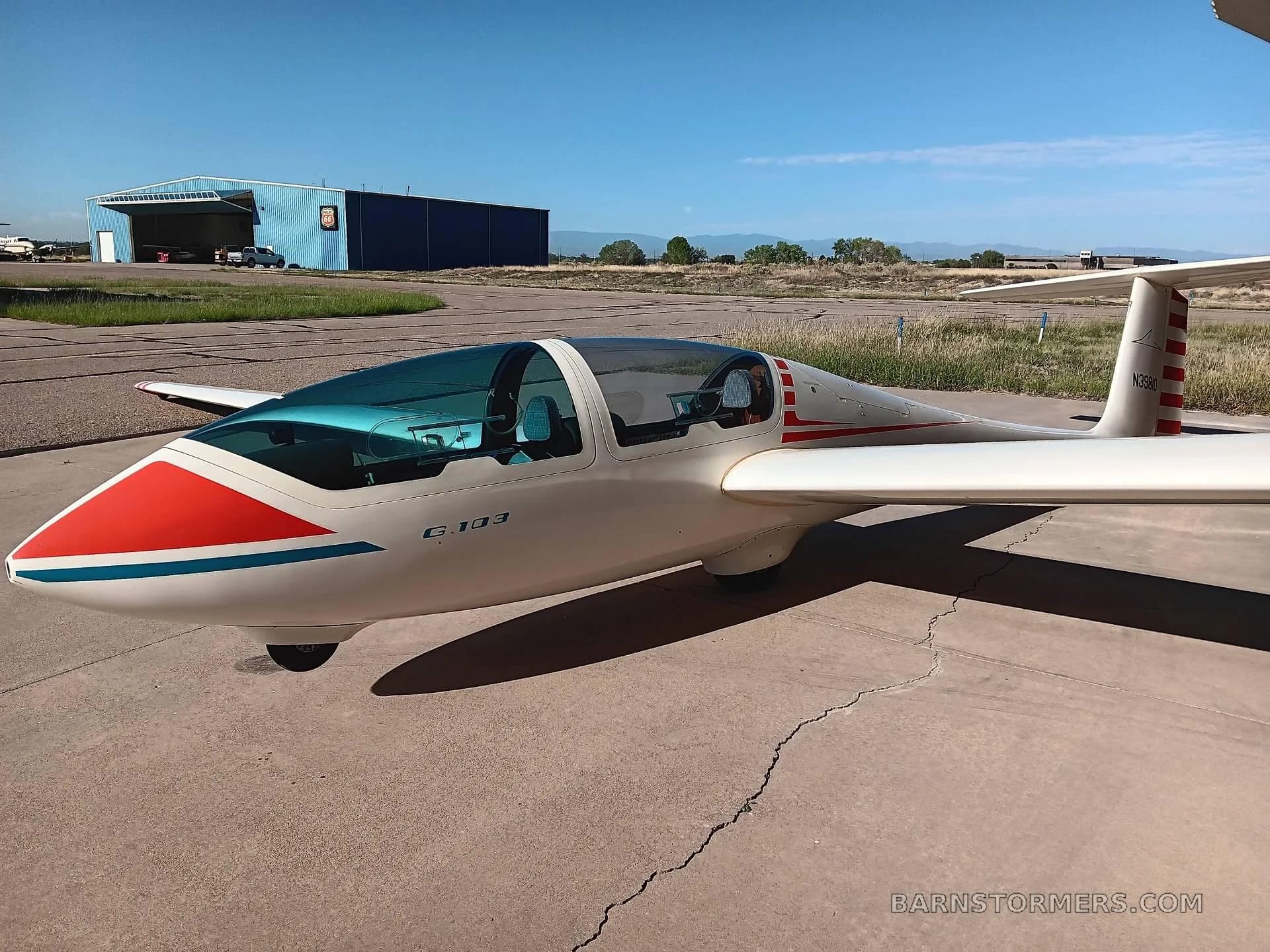 A white Grob G 103 glider with red and blue stripes on the nose, parked on an airfield tarmac with a blue hangar in the background.