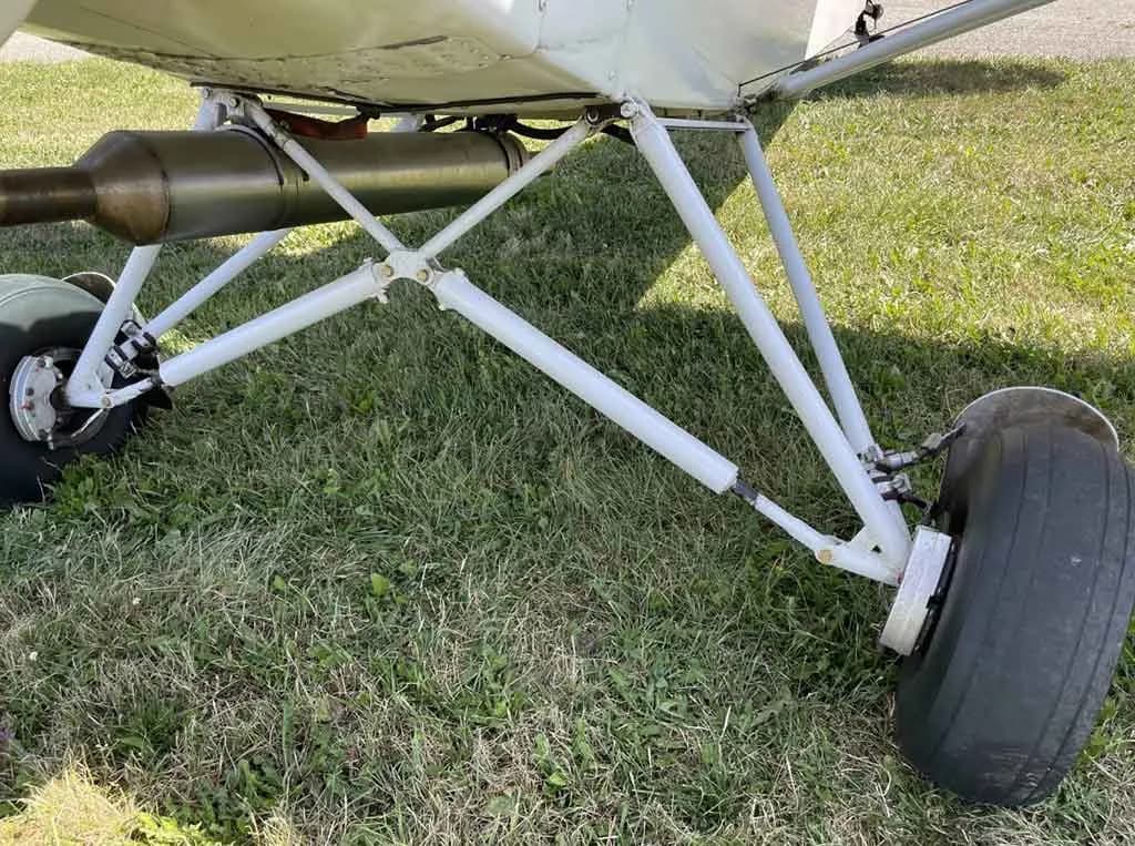 A close-up view of the white main landing gear struts and wheel of a light aircraft on grass.