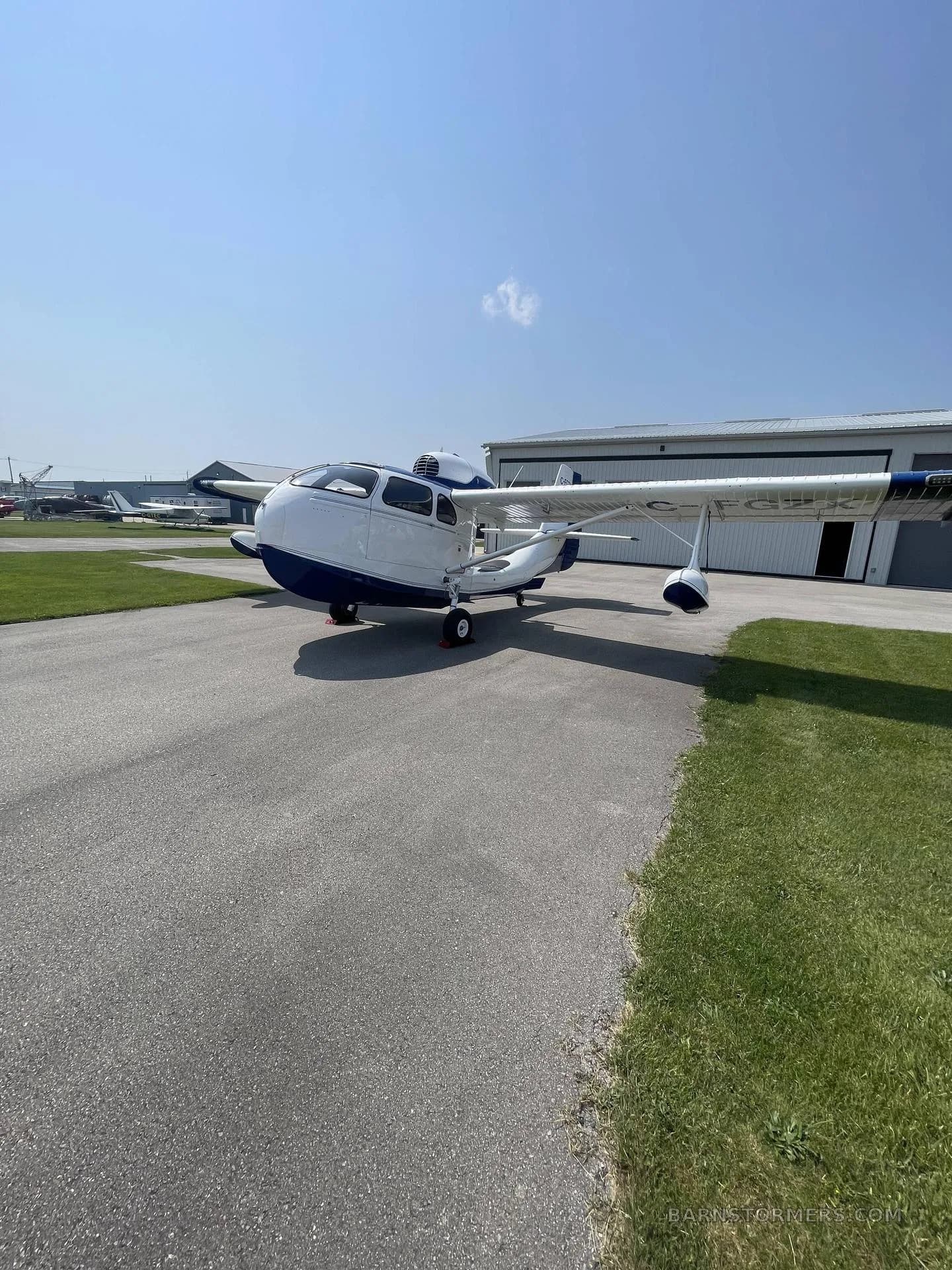 A white and blue amphibious aircraft with its wings extended, parked on an airfield tarmac with hangars in the background.