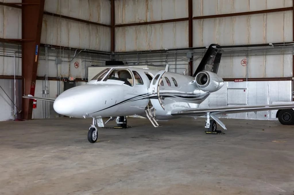 A white Cessna Citation M2 business jet with its main cabin door open is parked inside a hangar.