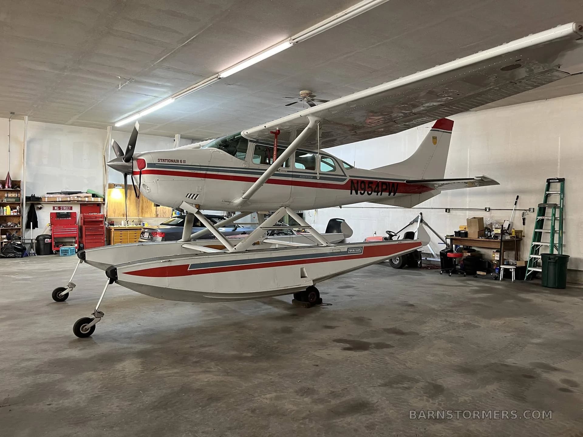 A white Cessna Stationair 6 II floatplane with red and blue stripes, registration N954PW, viewed from the left side in a hangar.