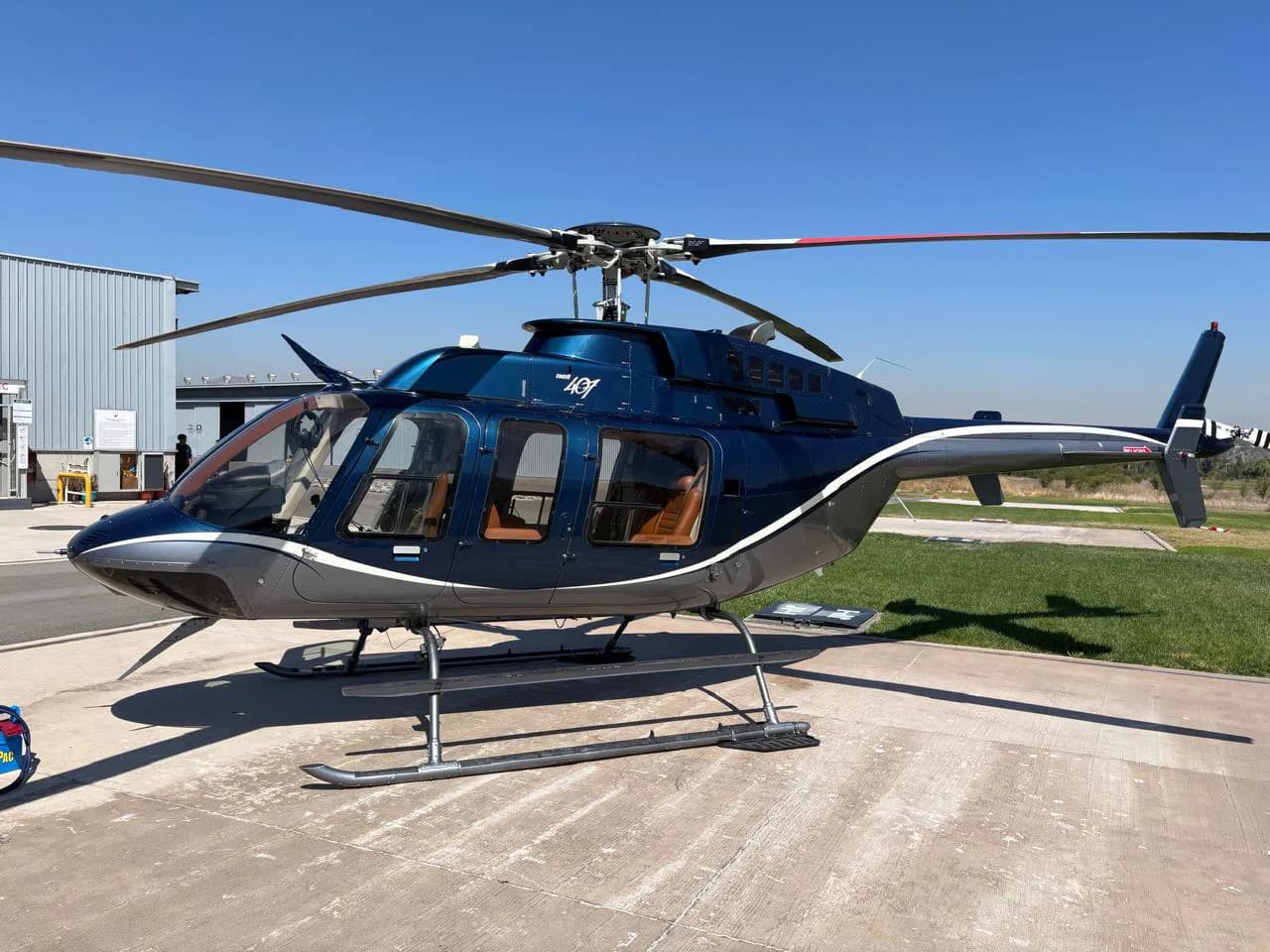 A dark blue and grey Bell 407 helicopter parked on a concrete pad next to a hangar on a sunny day.