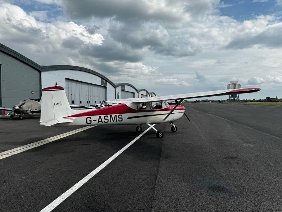 A white and red Cessna 150 G-ASMS parked on the tarmac, viewed from the rear-left, with hangars under a cloudy sky.