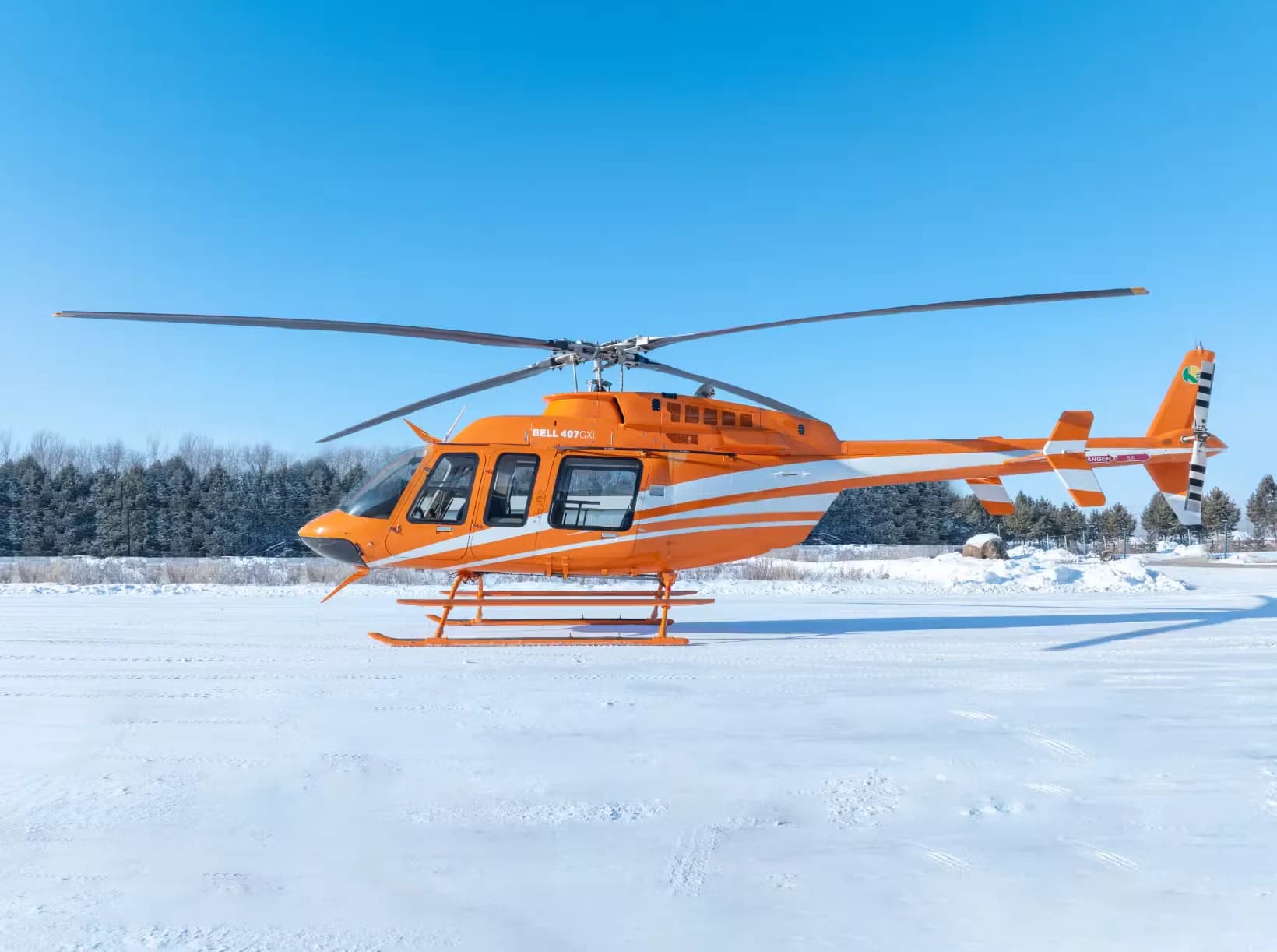 A Bell 407 helicopter with an orange and white paint scheme is parked on a snowy surface.