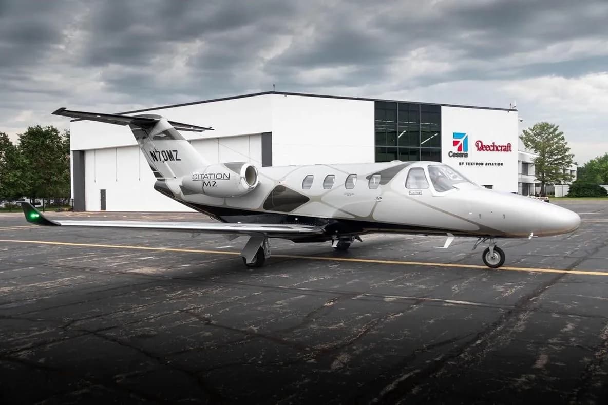A side view of a silver and black Cessna Citation M2 jet (N70MZ) parked on an asphalt ramp in front of a Textron Aviation hangar.