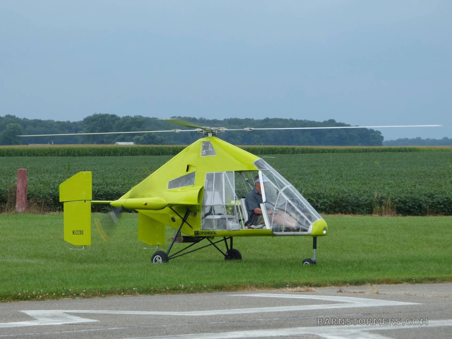 A bright green gyrocopter with a clear canopy and a pilot inside, parked on a grassy field next to a paved strip with cornfields in the background.