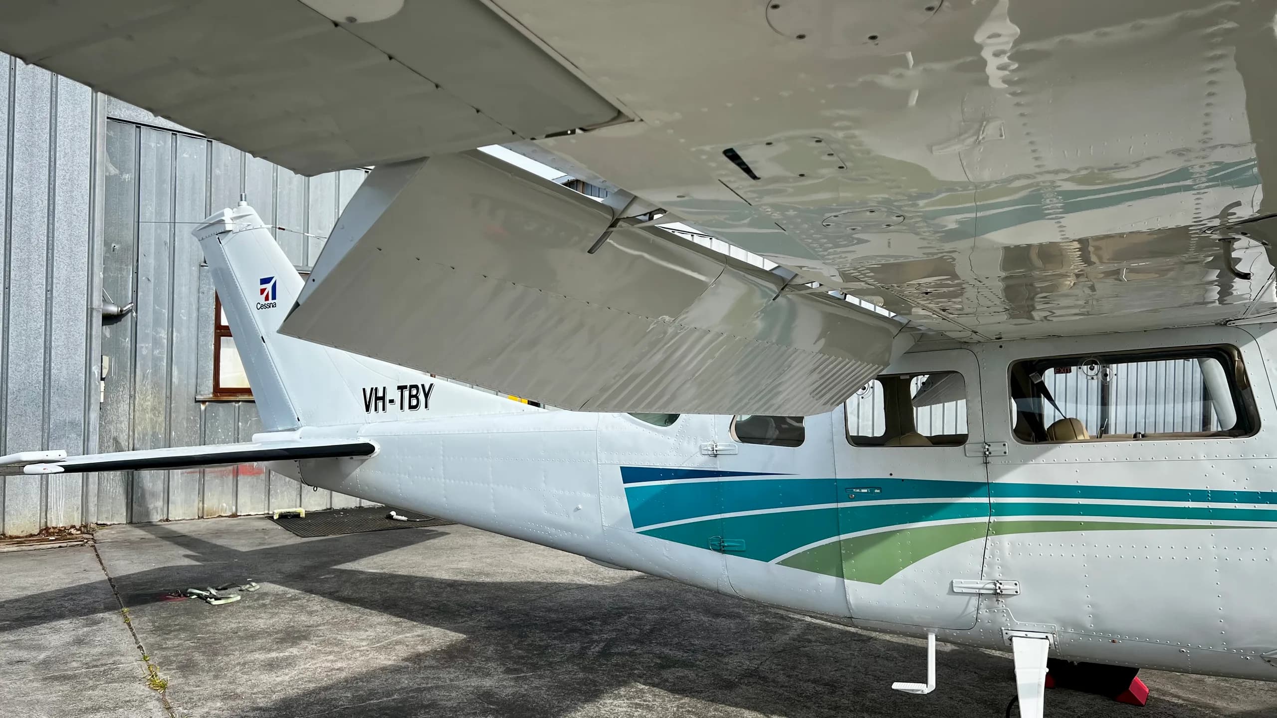 A close-up view of the tail and right wing of a white Cessna aircraft with blue and green stripes, showing the registration VH-TBY.