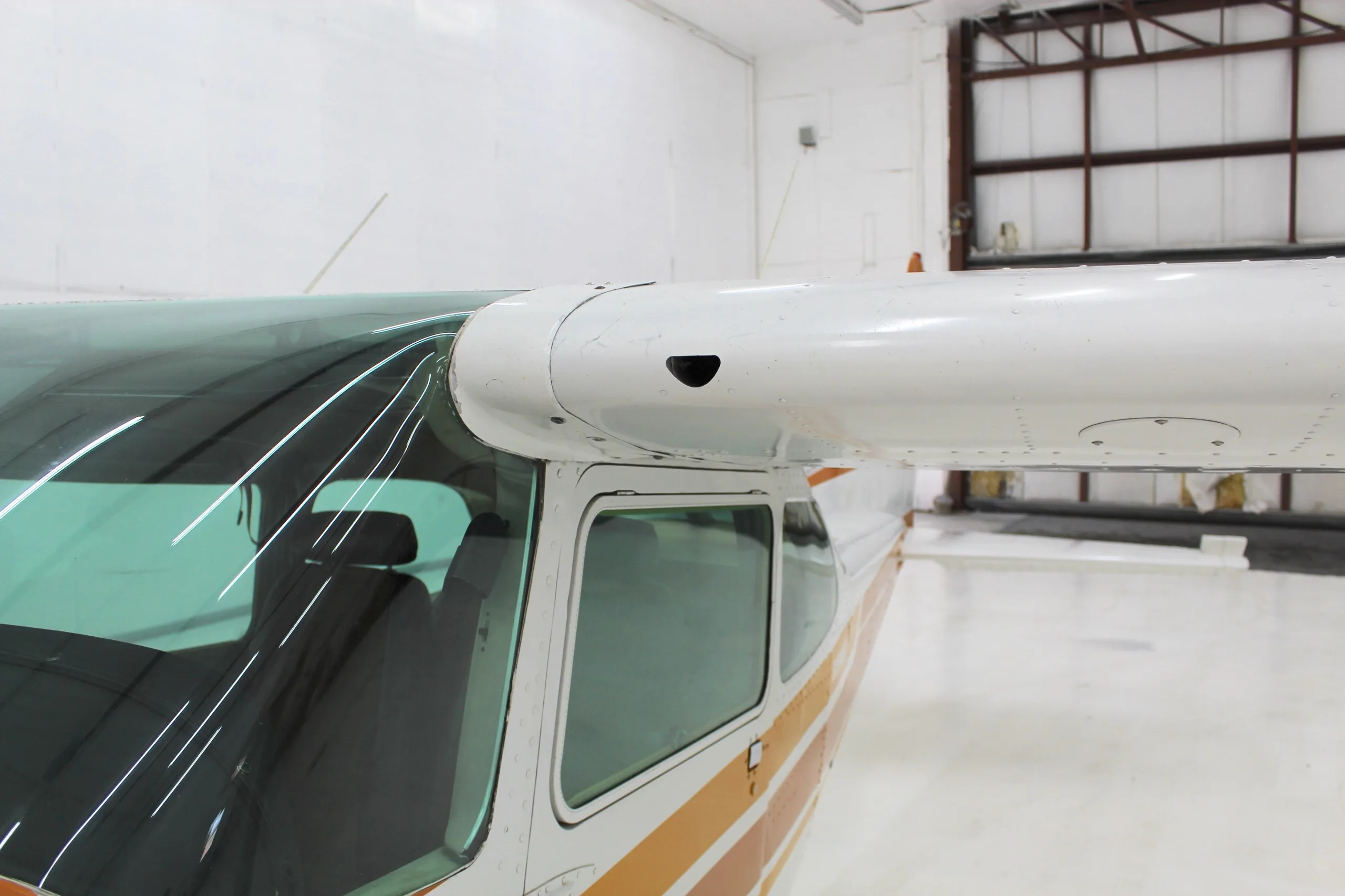 A close-up view of the wing root and cockpit windows of a white and orange Cessna Skyhawk II in a hangar.