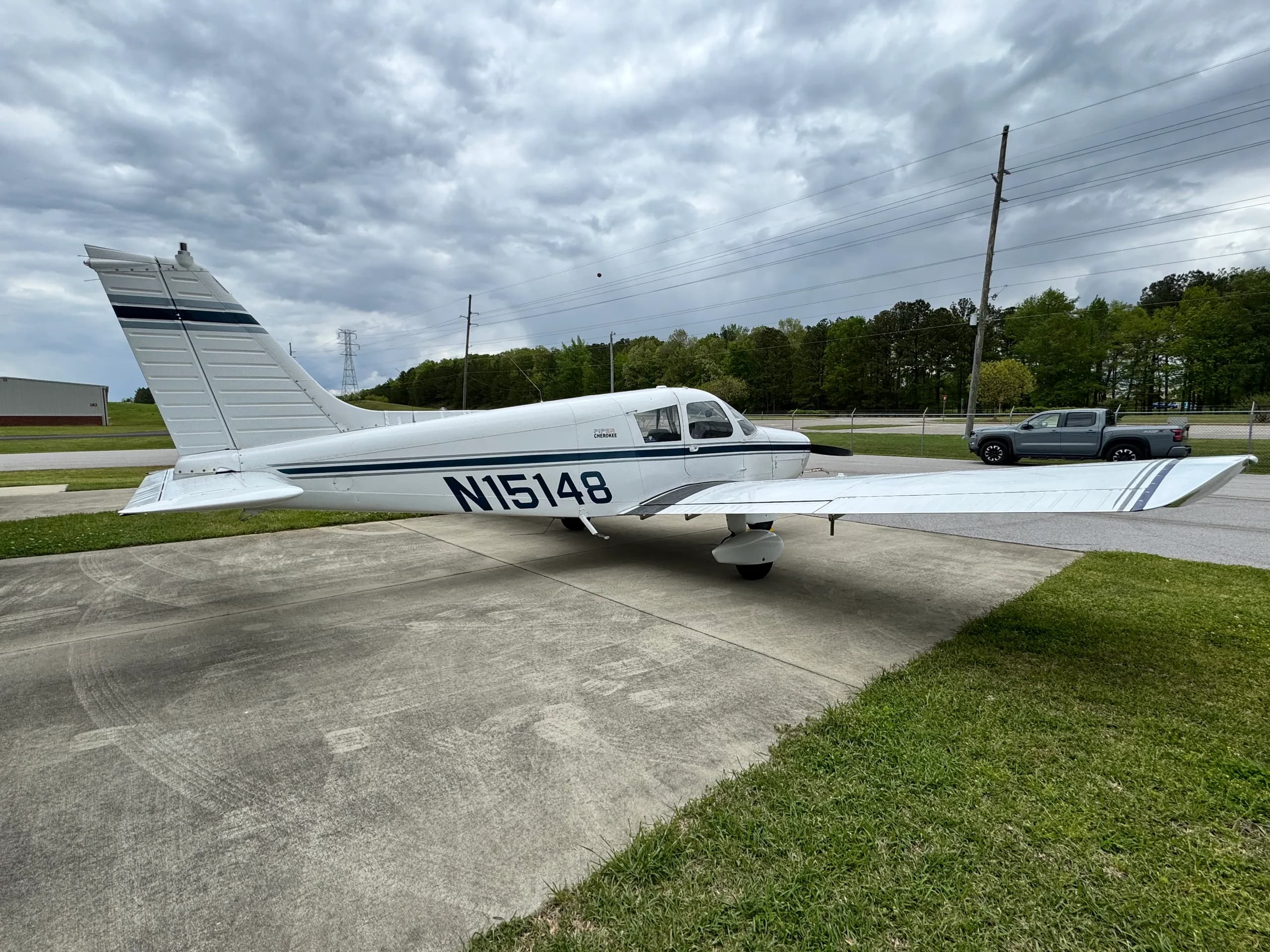 Side view of a white single-engine aircraft, N15148, with dark blue stripes, parked on concrete with trees and power lines in the background.