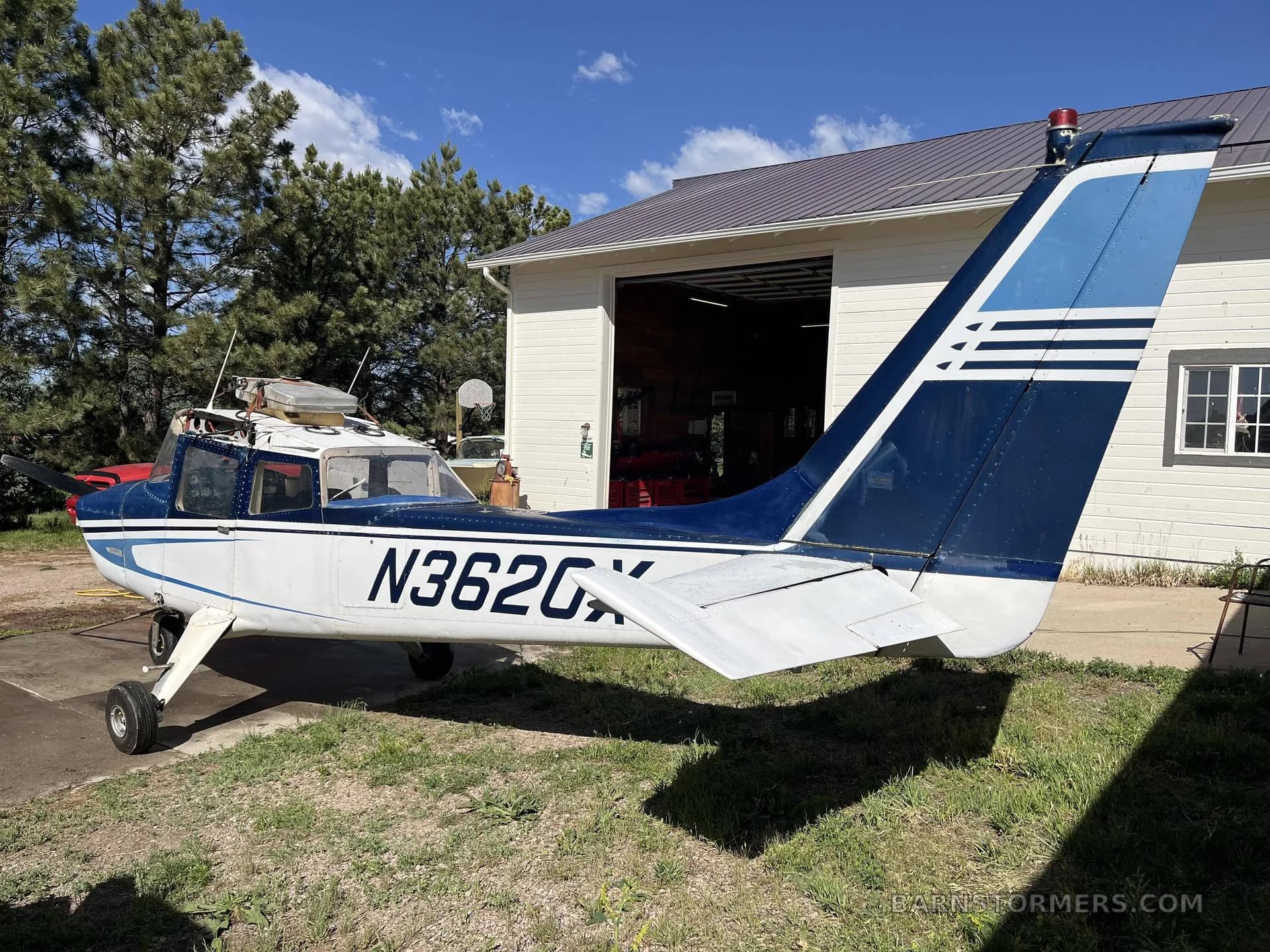 The rear-side view of a blue and white single-engine propeller aircraft (N3620X) parked on grass next to a hangar.