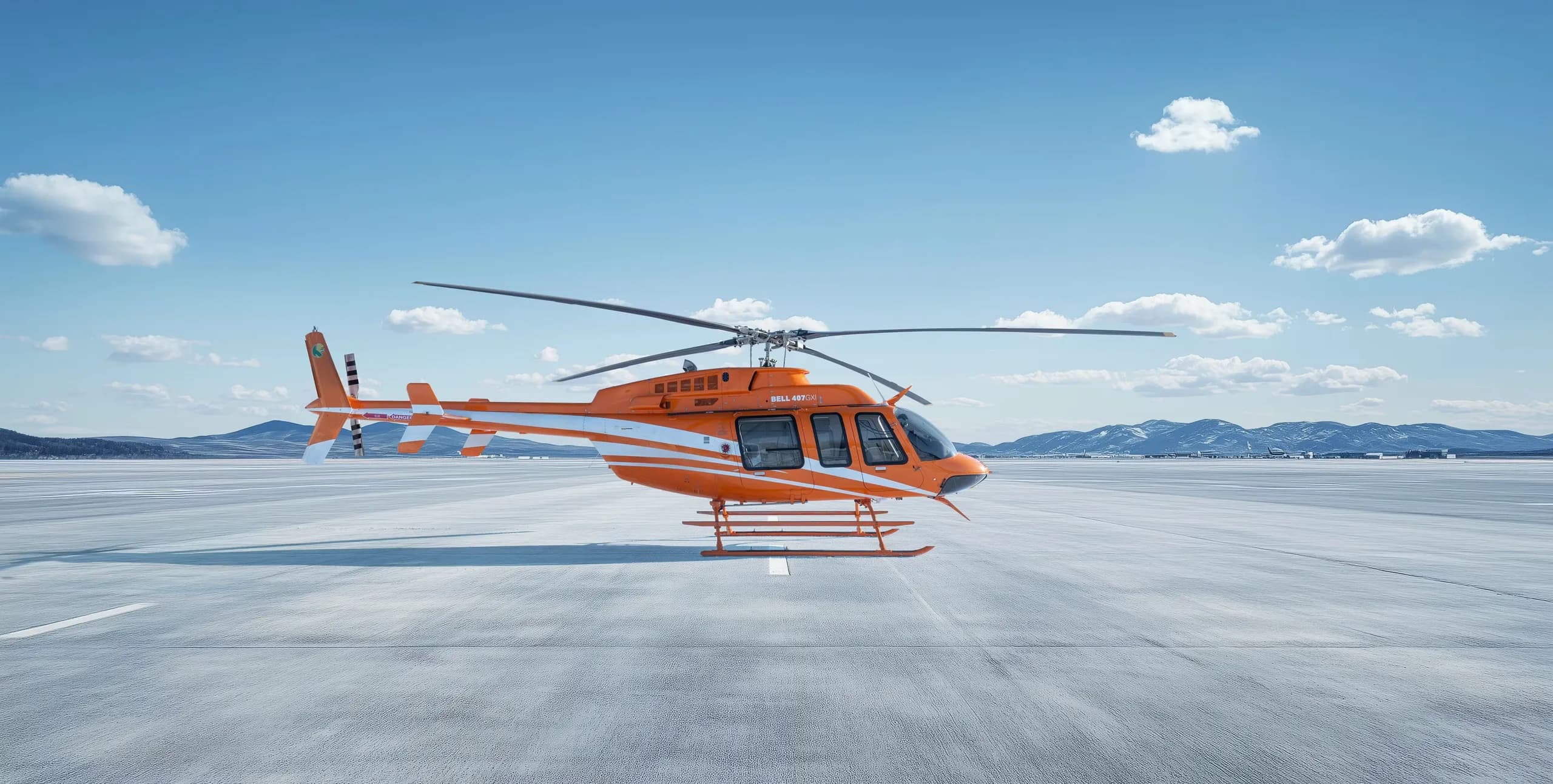 A Bell 407 helicopter with an orange and white paint scheme is parked on a concrete surface.