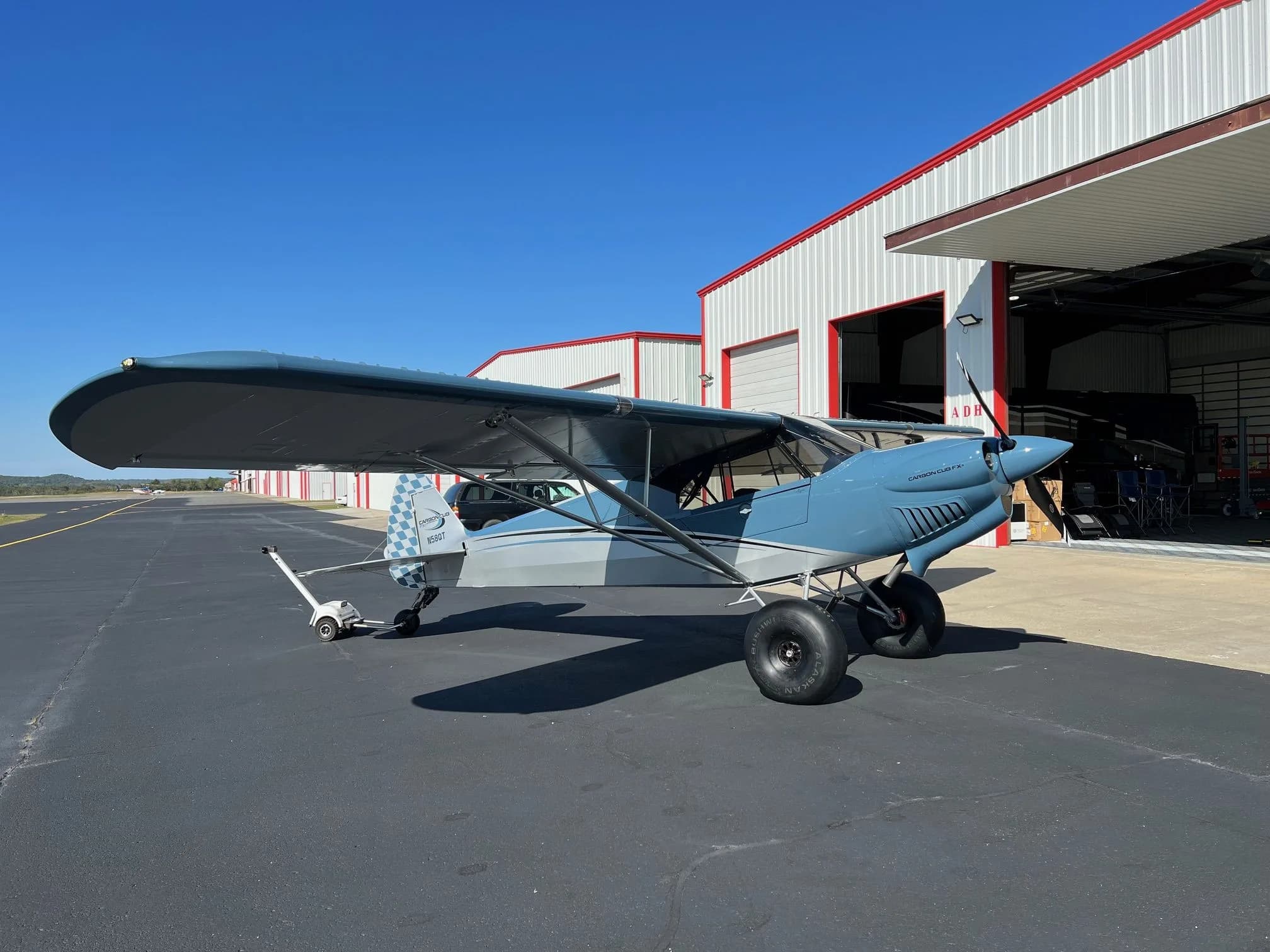 A blue and grey Carbon Cub FX-3 aircraft with registration N58QT parked on an airfield next to hangars.