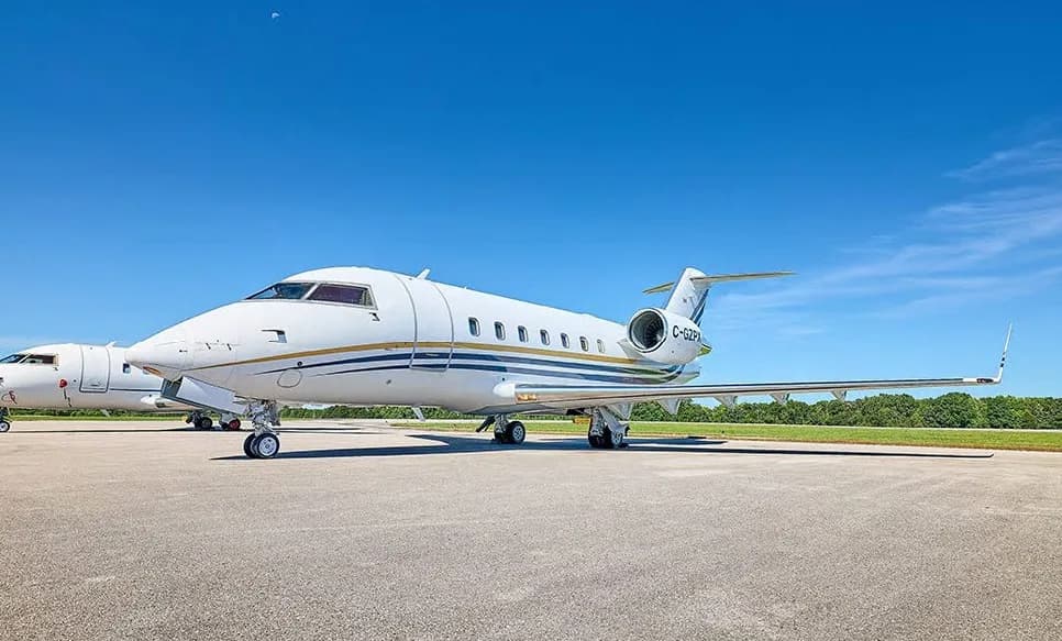 A white Bombardier Challenger business jet with blue and gold stripes is parked on an airport tarmac under a clear blue sky.