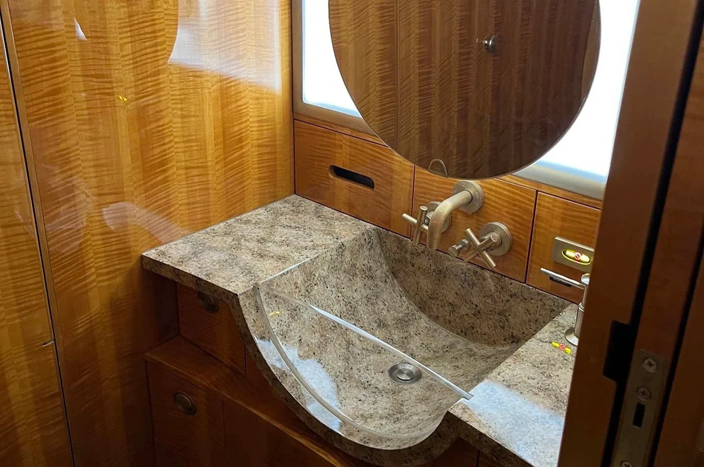 A modern aircraft lavatory with a unique glass and granite sink, wood paneling, and a round mirror.