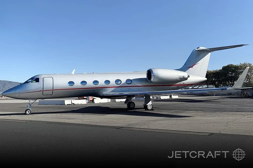 A silver Gulfstream business jet with a red stripe is parked on an asphalt tarmac under a clear blue sky.