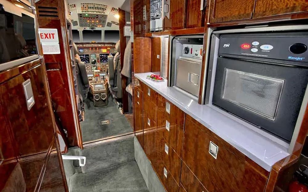 The galley area of a business jet with dark wood cabinetry, a microwave, and a view towards the cockpit.
