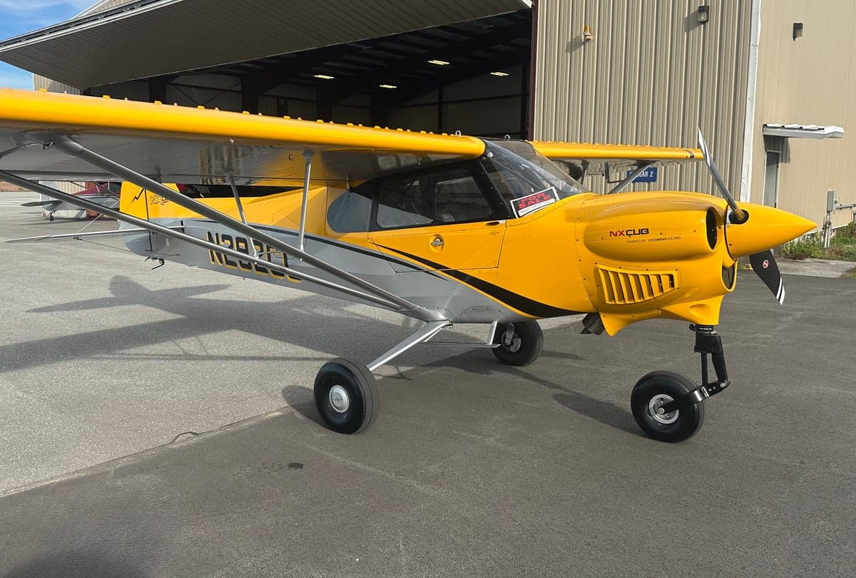 A yellow and gray NX Cub aircraft, N292CC, parked in front of an open hangar.