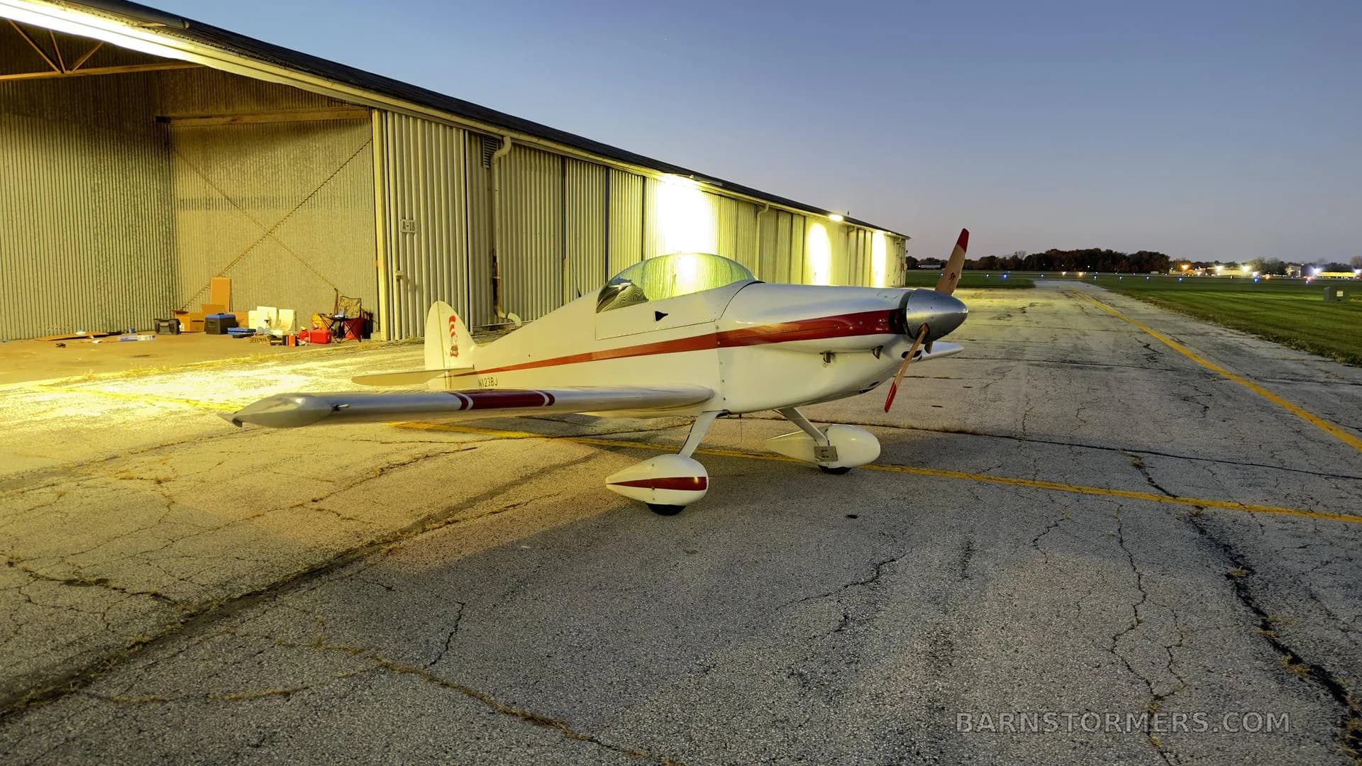 A white and red single-engine aircraft with a propeller, parked on an asphalt apron next to a hangar at dusk, illuminated by hangar lights.