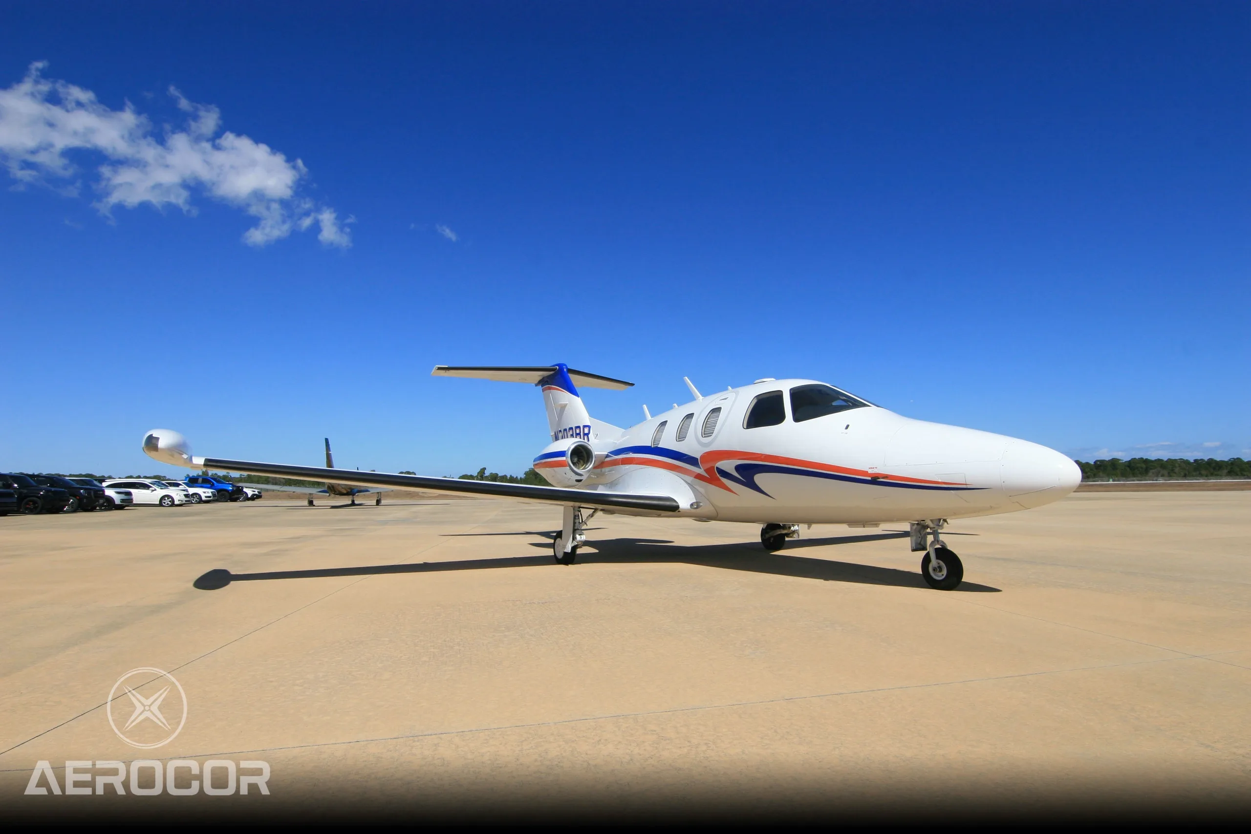 A white Eclipse 500 jet with blue and orange stripes, tail number N703BR, parked on an airfield with other aircraft in the background.