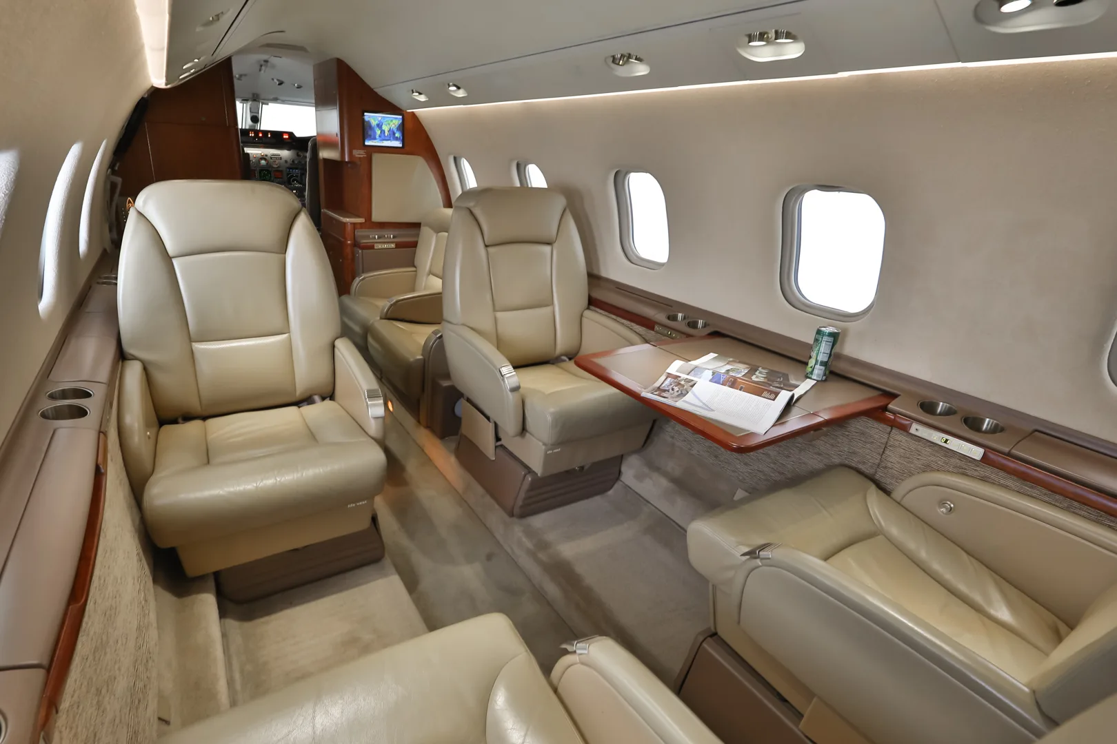 An angled view of the business jet's cabin, showing beige leather seats, a fold-out table with a magazine, and large windows.