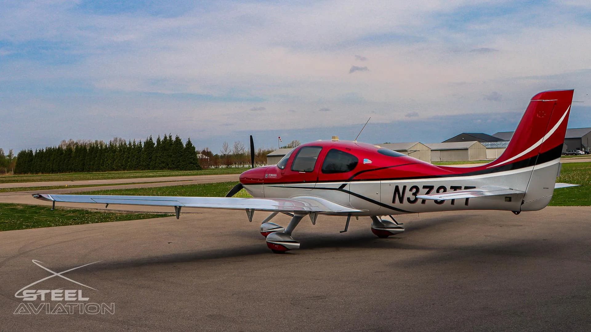 A red and white Cirrus SR22 aircraft with tail number N378TT is parked on an apron, viewed from the left side with hangars and trees in the background.