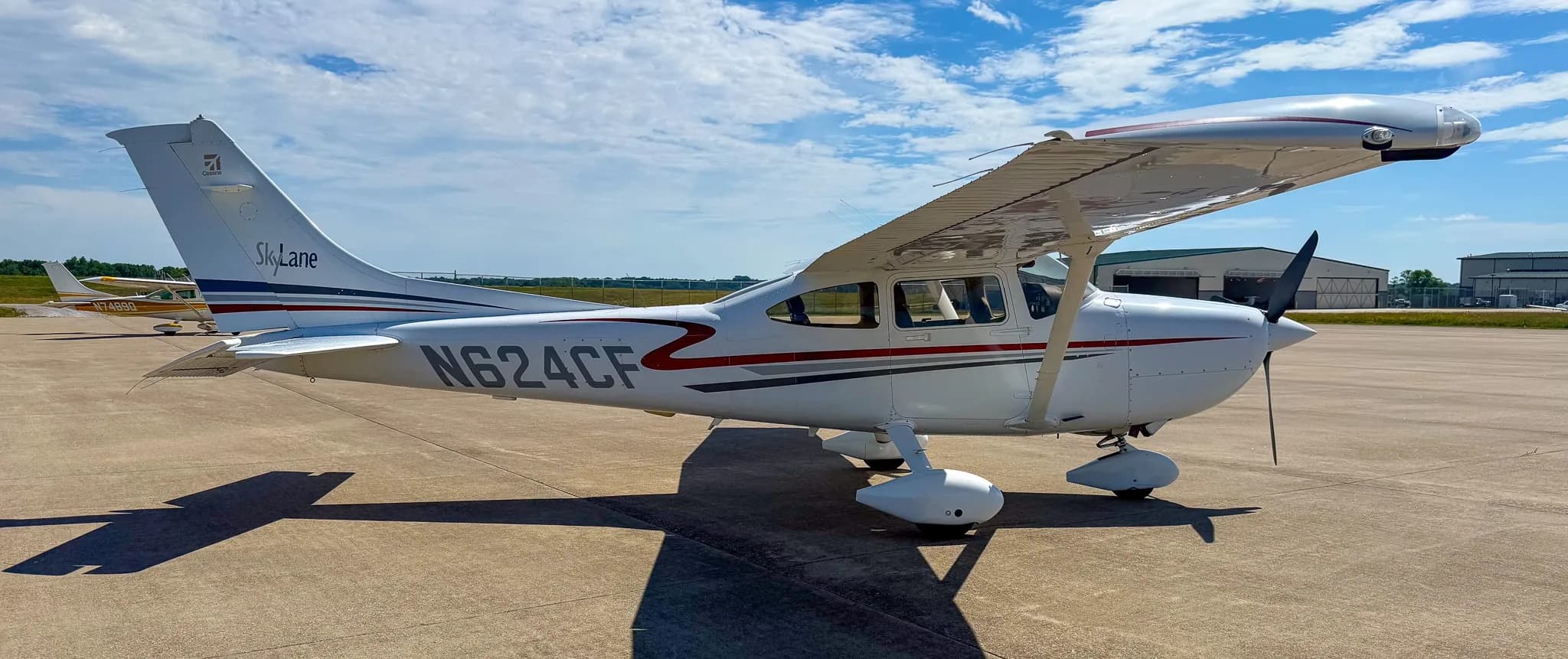 A white Cessna 182 Skylane with red and gray stripes, registration N624CF, parked on an asphalt ramp near a hangar.
