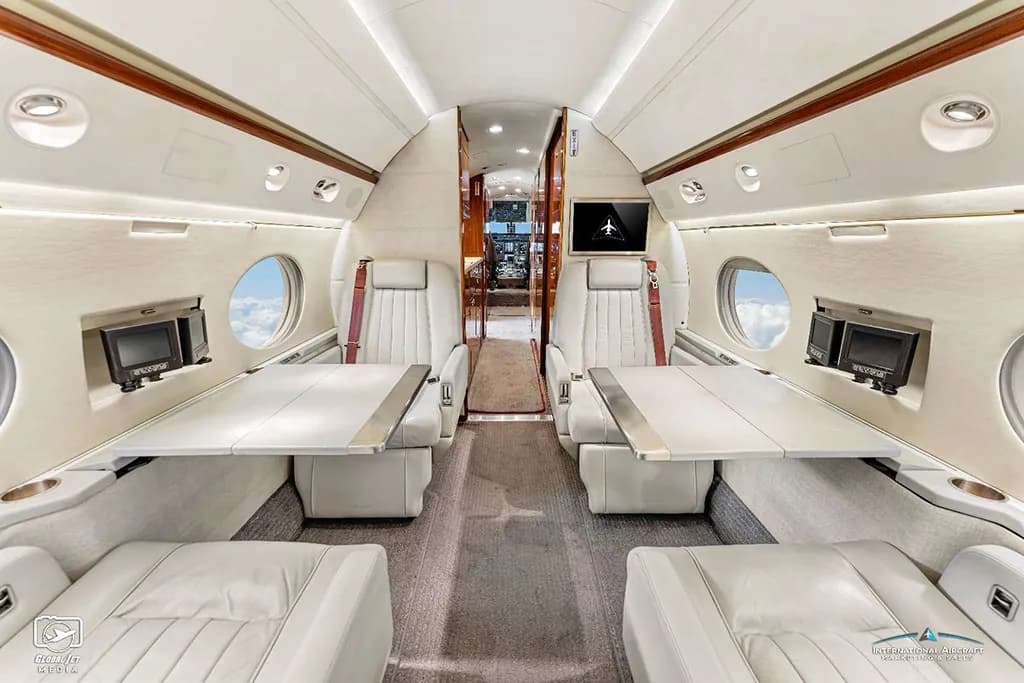 The mid-cabin interior of a Gulfstream G550, showing light-colored seats with deployed tables and entertainment screens.