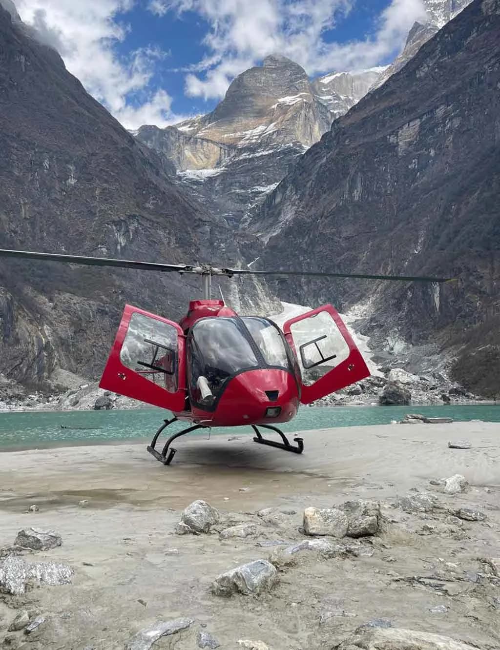 A red helicopter with open doors is parked on a rocky shore by a turquoise lake, surrounded by towering mountains.