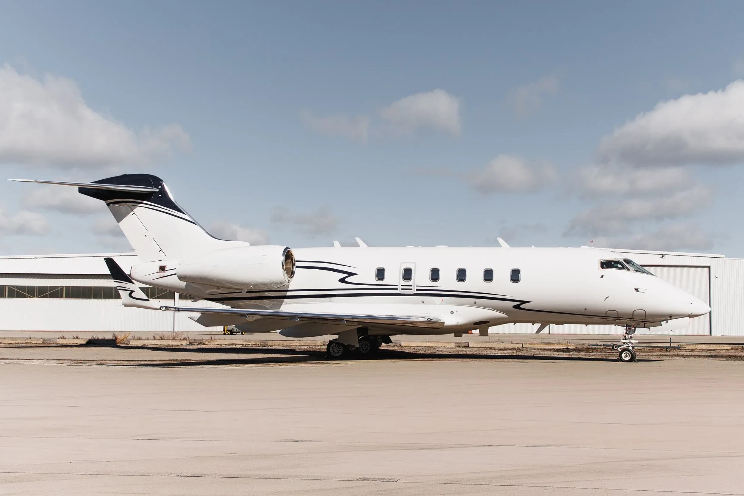 A white business jet with black and blue stripes on the tail and fuselage parked on an tarmac in front of a hangar under a partly cloudy sky.