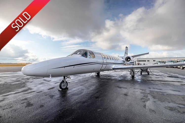 A white business jet with a black stripe is parked on a wet tarmac under a cloudy sky.