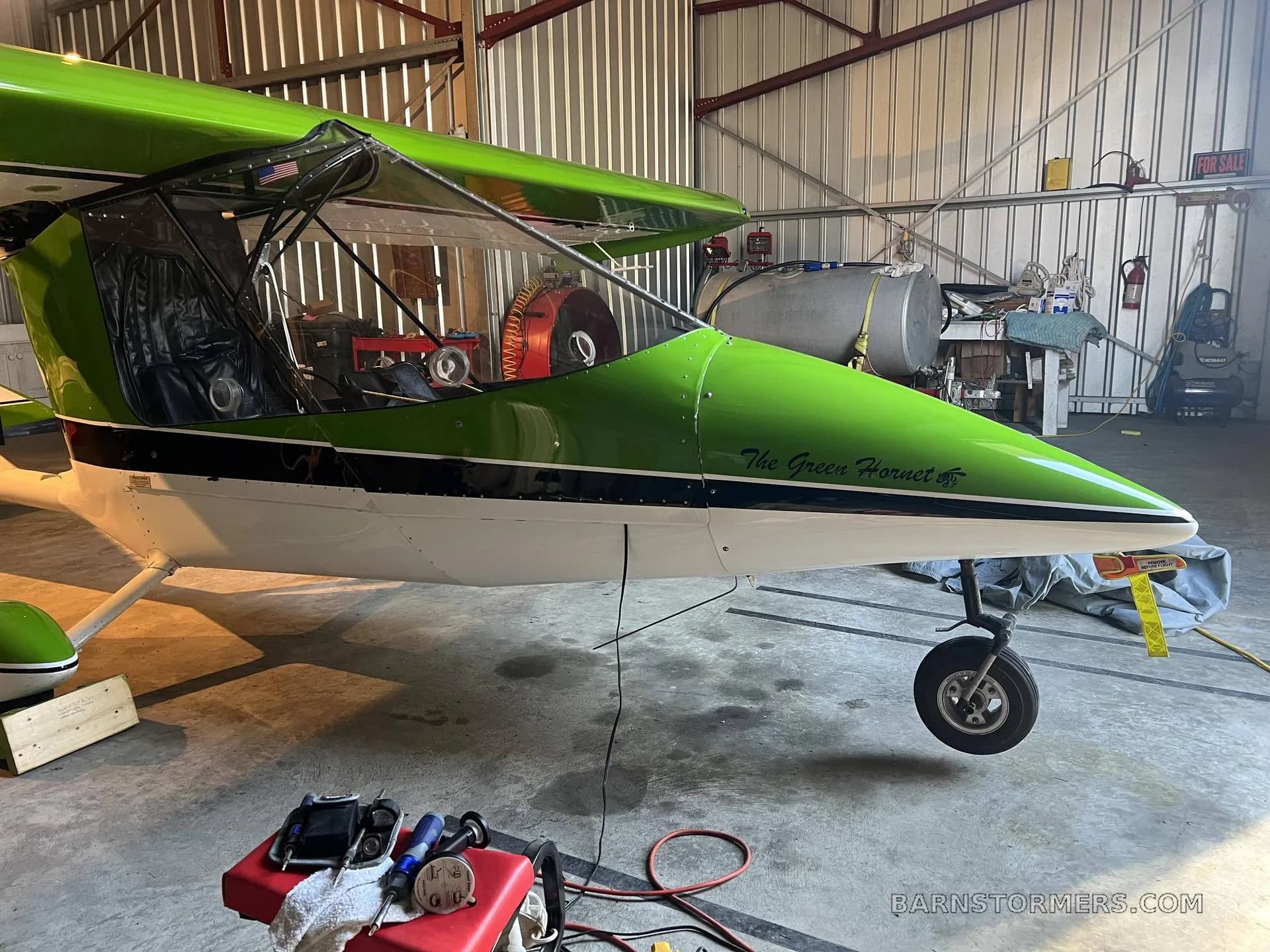 The front section of a green and white ultralight aircraft, named "The Green Hornet," is shown inside a hangar.