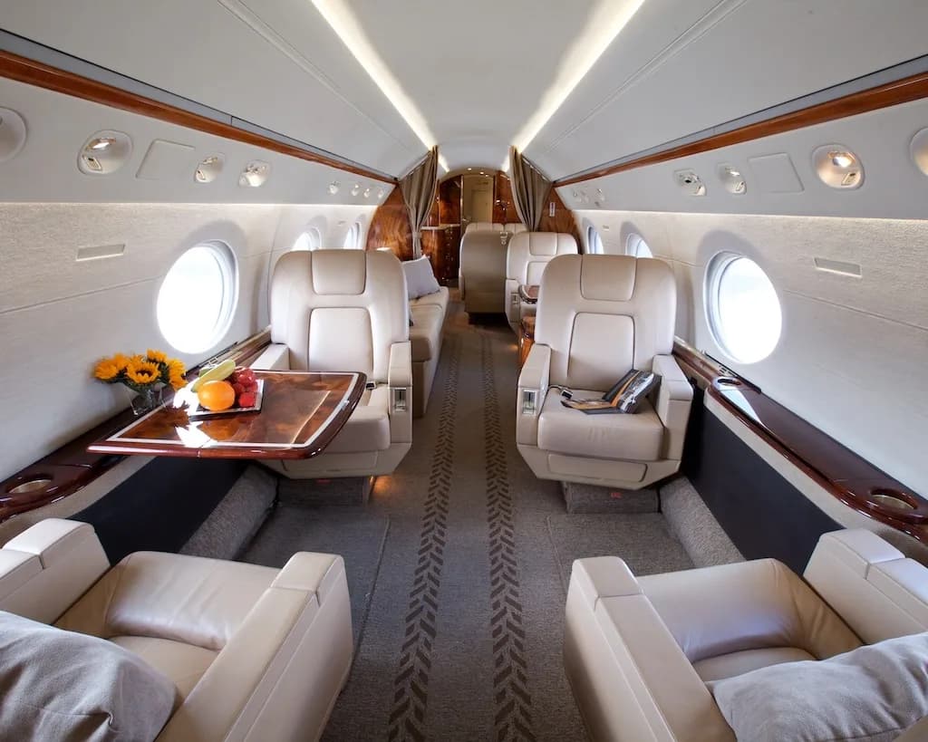 The luxurious interior cabin of a private jet, featuring beige leather seats, a wooden table with fruit, and large windows.