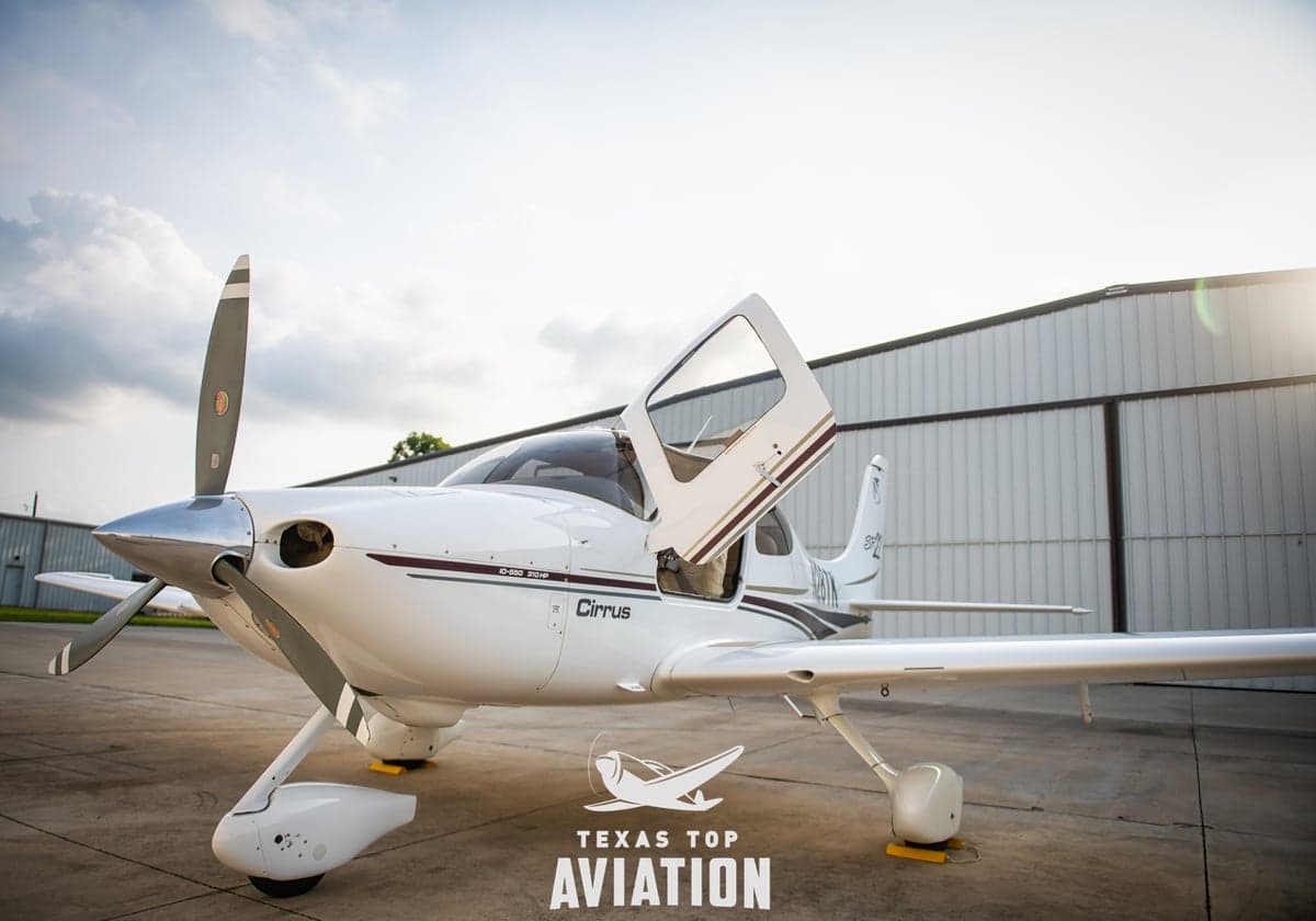 A Cirrus aircraft is parked on the tarmac with its doors open, featuring a white and brown paint scheme.