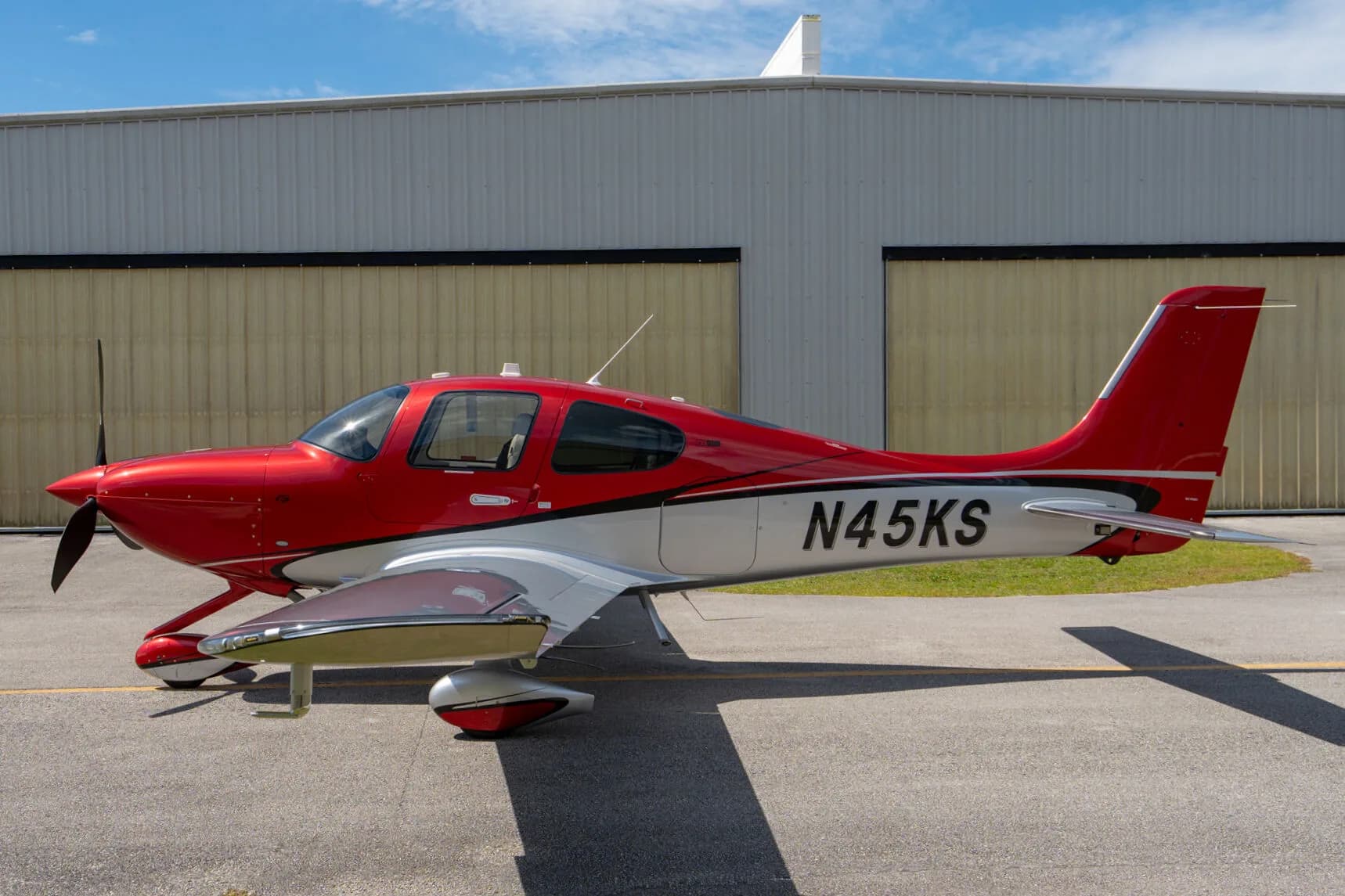 Side profile of a red and silver Cirrus SR22 GTS (N45KS) parked on an airfield next to a hangar.
