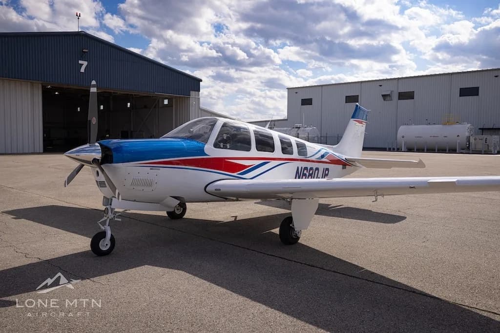 A front-side view of a white, blue, and red Beechcraft Bonanza N680JP parked on an airfield in front of a hangar.