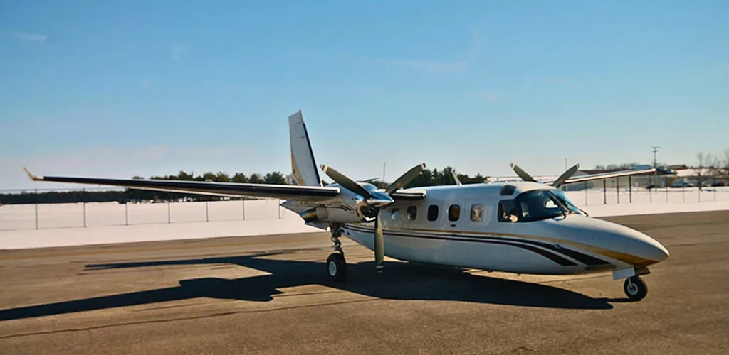 A white twin-engine turboprop aircraft with gold and maroon stripes parked on an airfield with snow-covered ground in the distance.