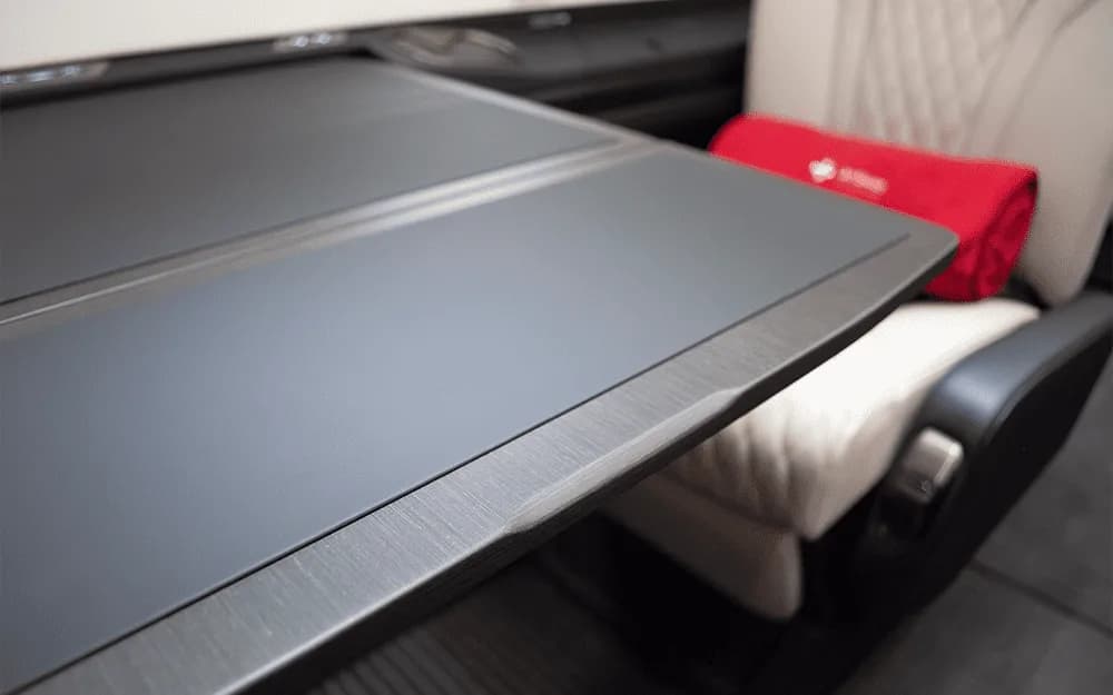 A close-up view of a dark grey fold-out table and a white quilted leather seat in the Pilatus PC-12 NGX cabin.