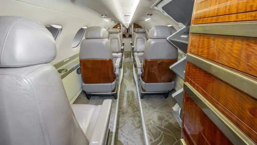 The interior cabin of a private jet, featuring rows of light grey leather seats with wooden accents and a patterned carpet.