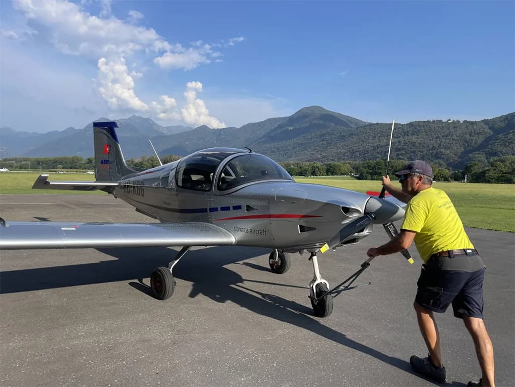 A silver Sonaca 200 aircraft with red and blue accents is being pushed by a person on a tarmac with mountains in the background.
