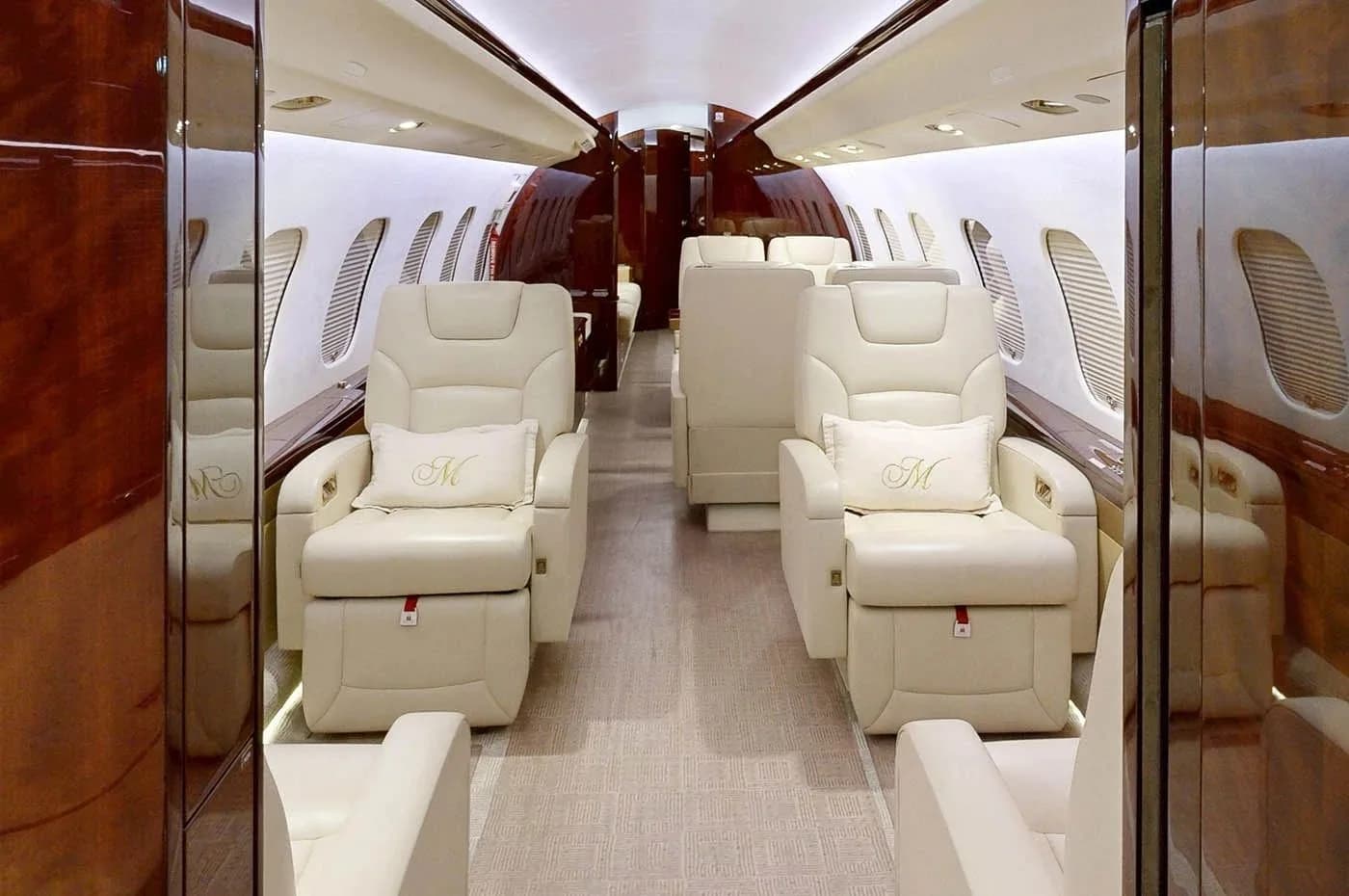 The luxurious cabin interior of a private jet, featuring cream-colored leather seats with "M" monogrammed pillows and polished dark wood accents.