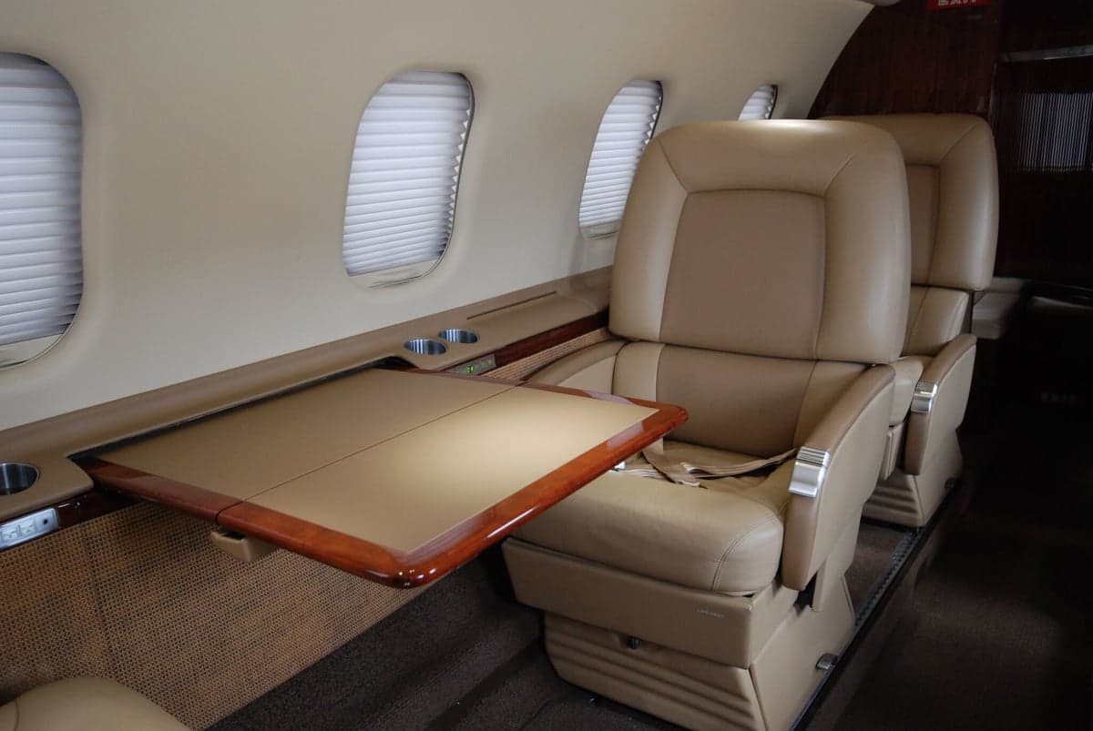 Beige leather passenger seats in a business jet cabin with a polished wood fold-out table extended from the sidewall.