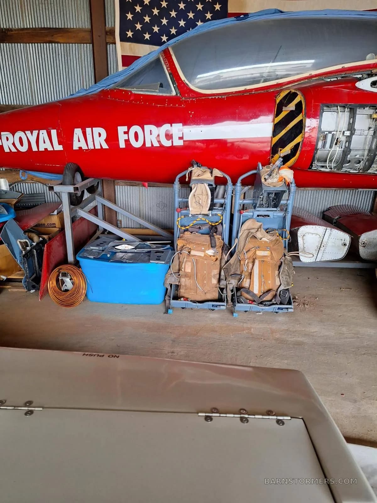 The front section of a red jet aircraft with 'ROYAL AIR FORCE' text, partially disassembled in a hangar with ejection seats in the foreground.